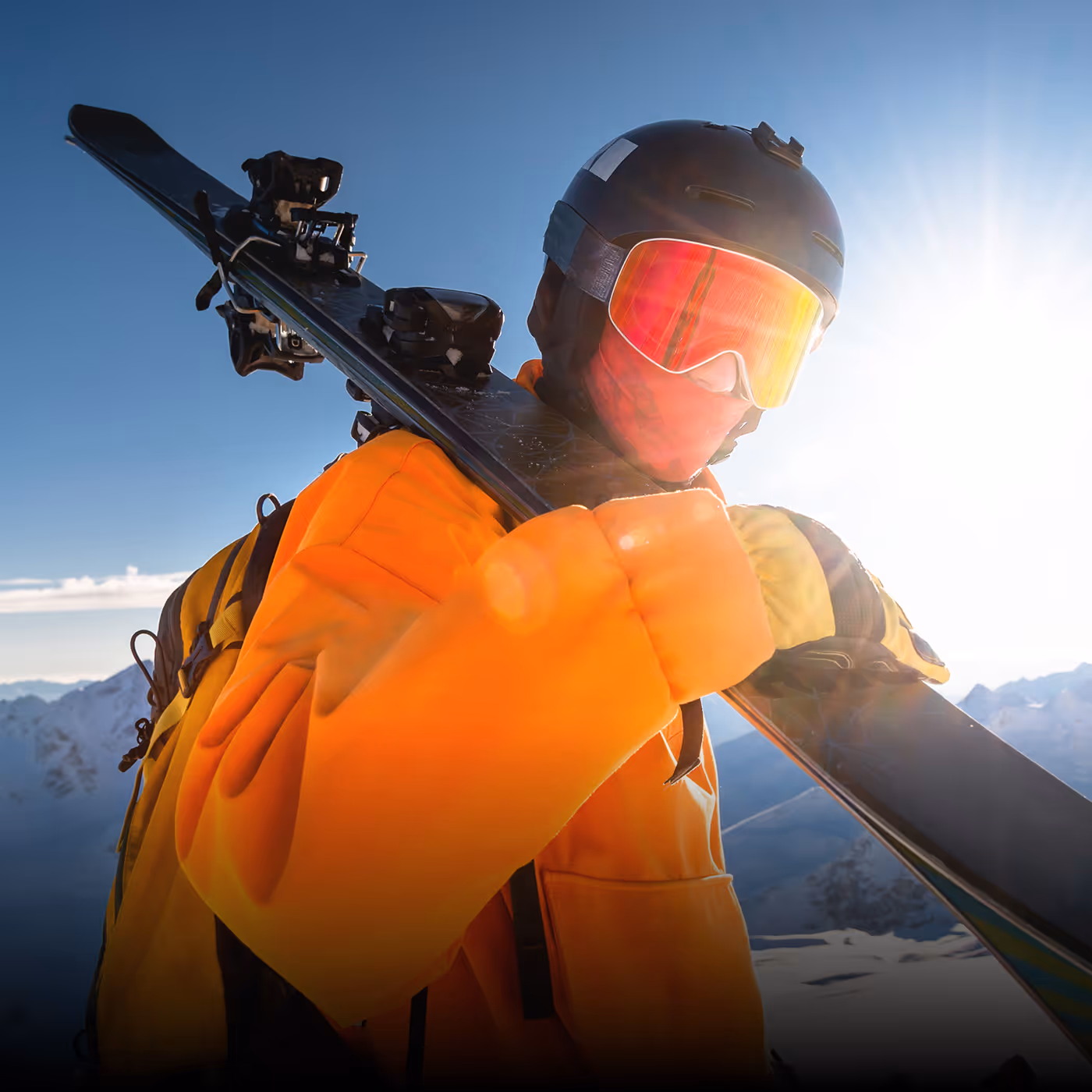 Skier wearing an orange jacket, helmet, and reflective goggles carrying skis over the shoulder with snowy mountains in the background for Pioneer Ridge brand