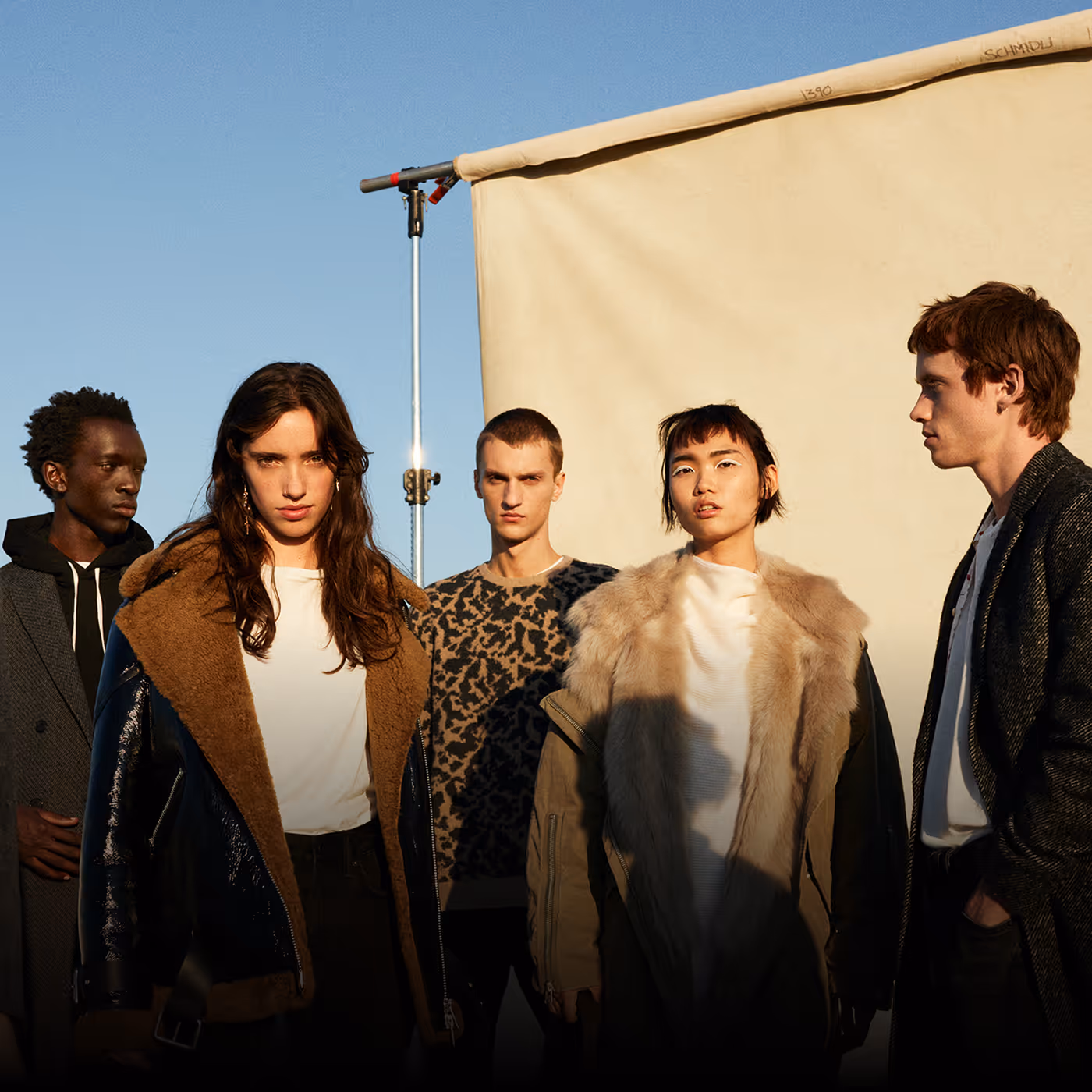 Five diverse young adults wearing stylish winter coats and jackets standing outdoors against a beige backdrop under a clear blue sky for AllSaints brand
