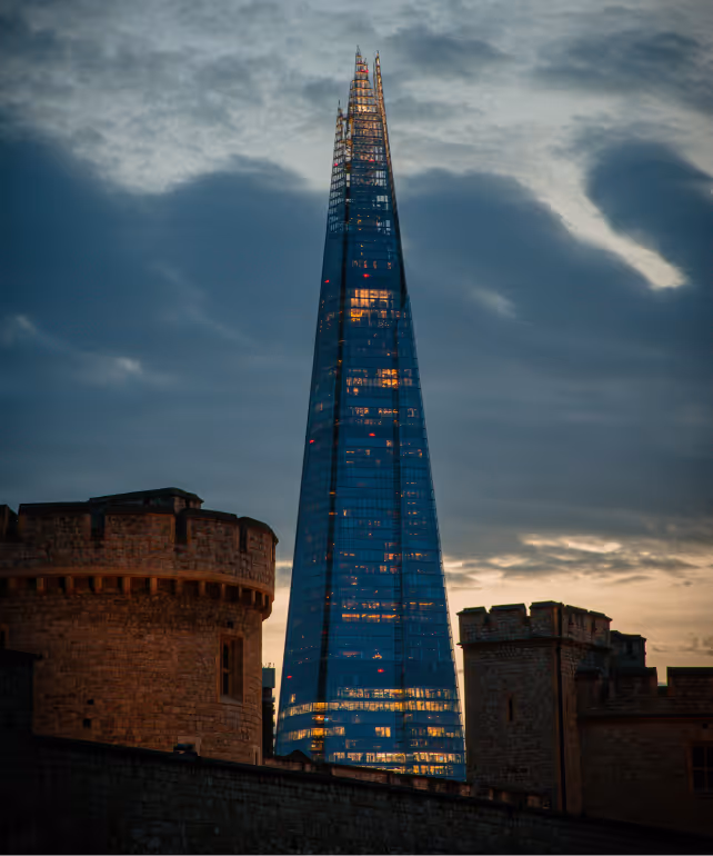 The Shard skyscraper illuminated at dusk with dark clouds in the sky, framed by historic stone towers in the foreground