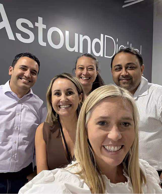 Group of five smiling adults taking a selfie in front of a gray wall with Astound Digital logo