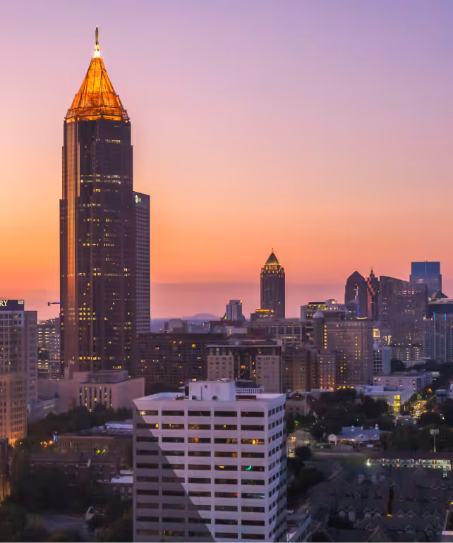 City skyline at sunset with a prominent skyscraper topped with golden lights against a colorful purple and orange sky