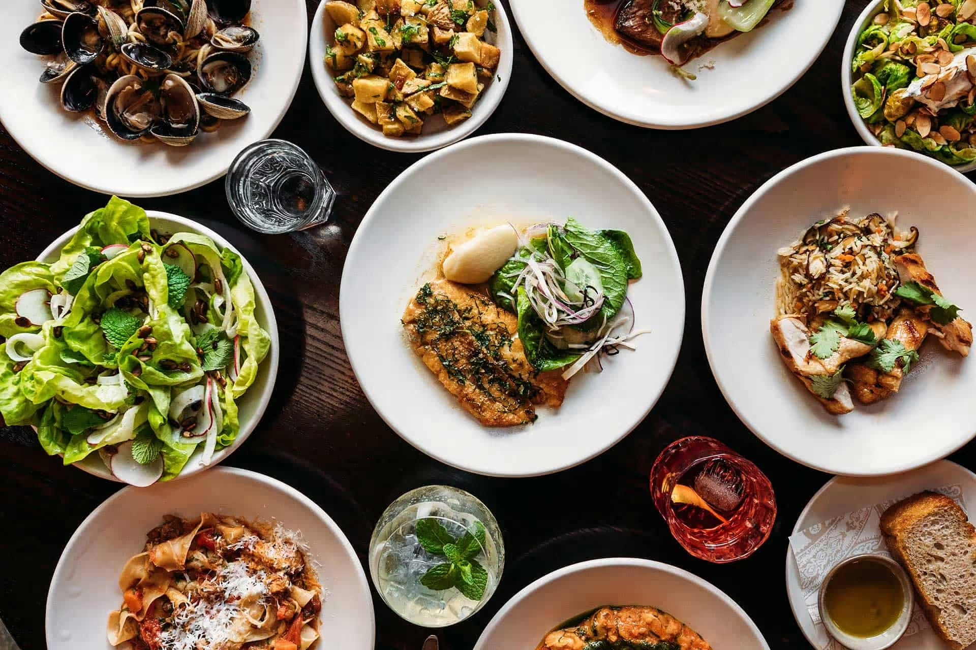 Top-down view of a variety of plated dishes including salad, pasta, seafood, grilled chicken, and drinks arranged on a dark wooden table.