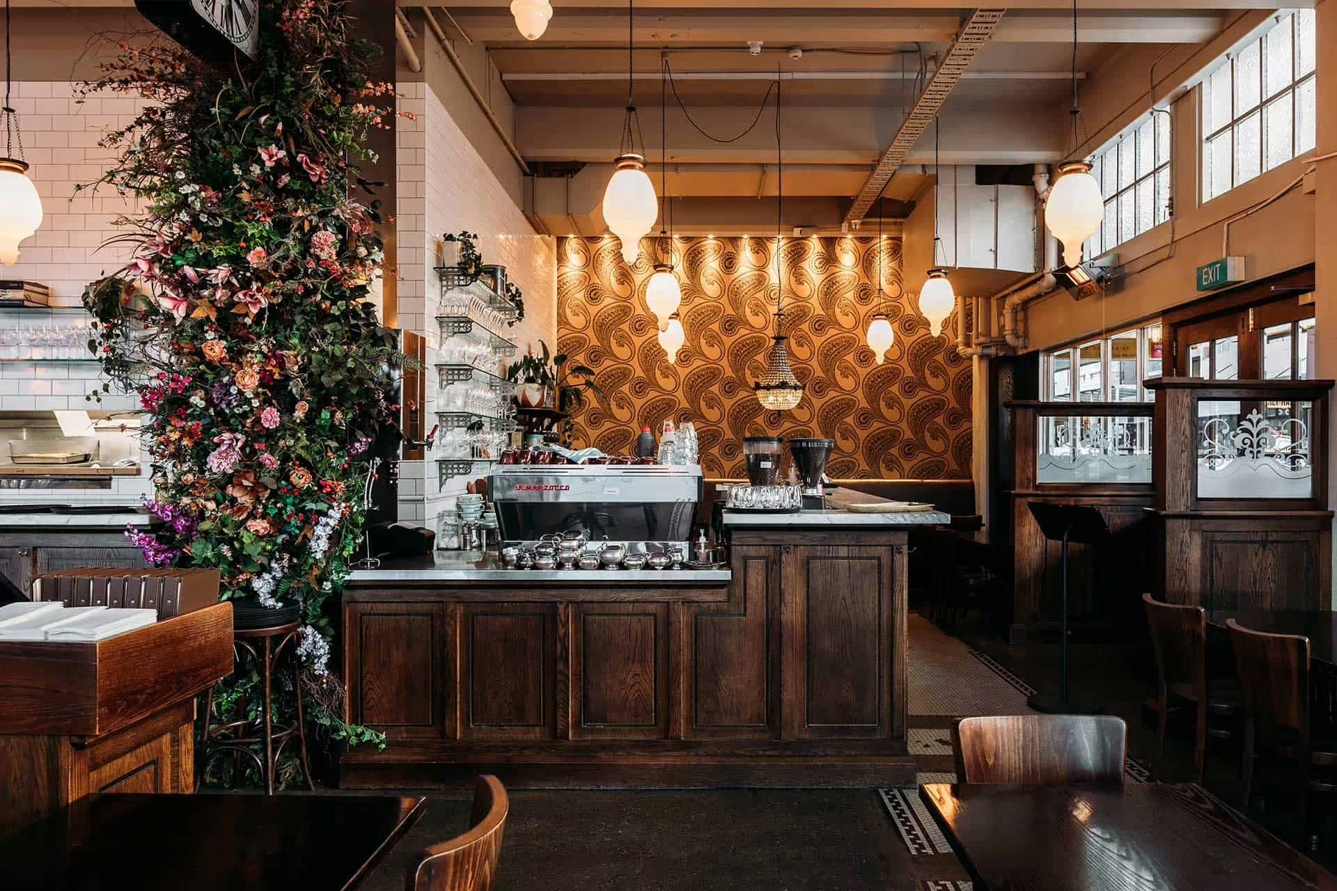 Cozy coffee shop interior with dark wood counters, hanging lights, a large floral arrangement, and patterned wallpaper.