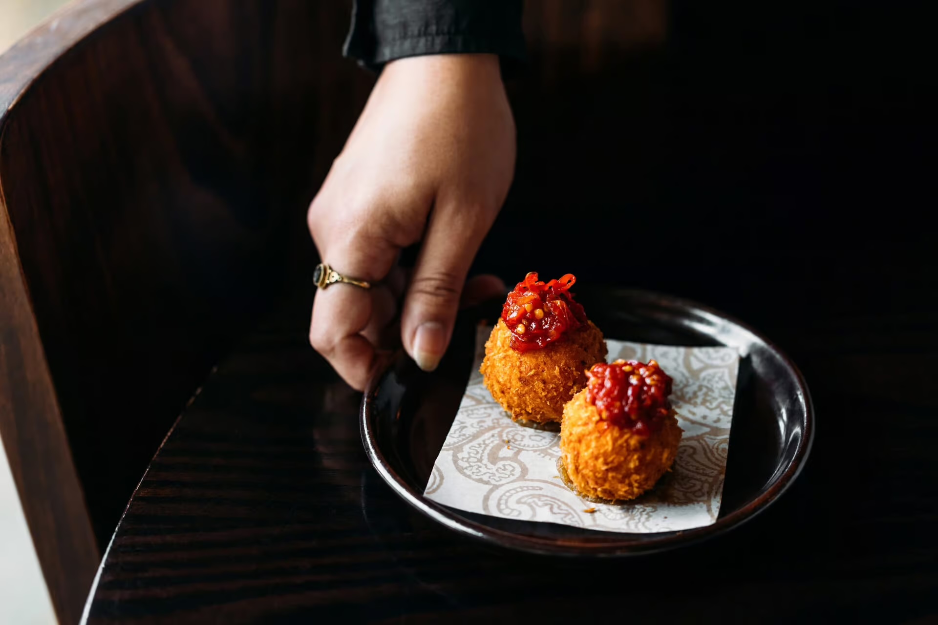 Hand holding a black plate with two crispy fried balls topped with red sauce on patterned paper.