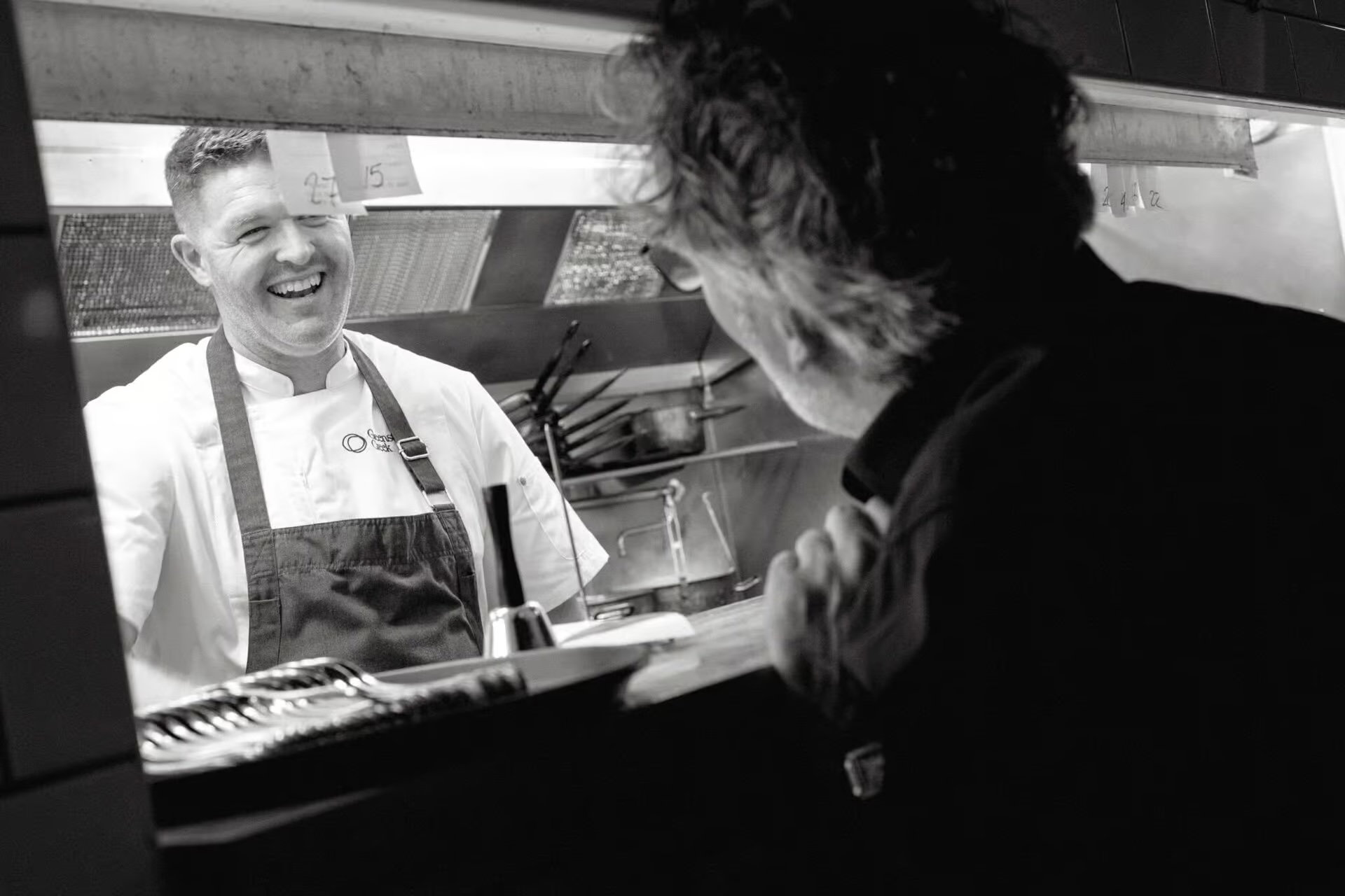 Smiling chef wearing apron conversing with a customer at kitchen pass in a restaurant.