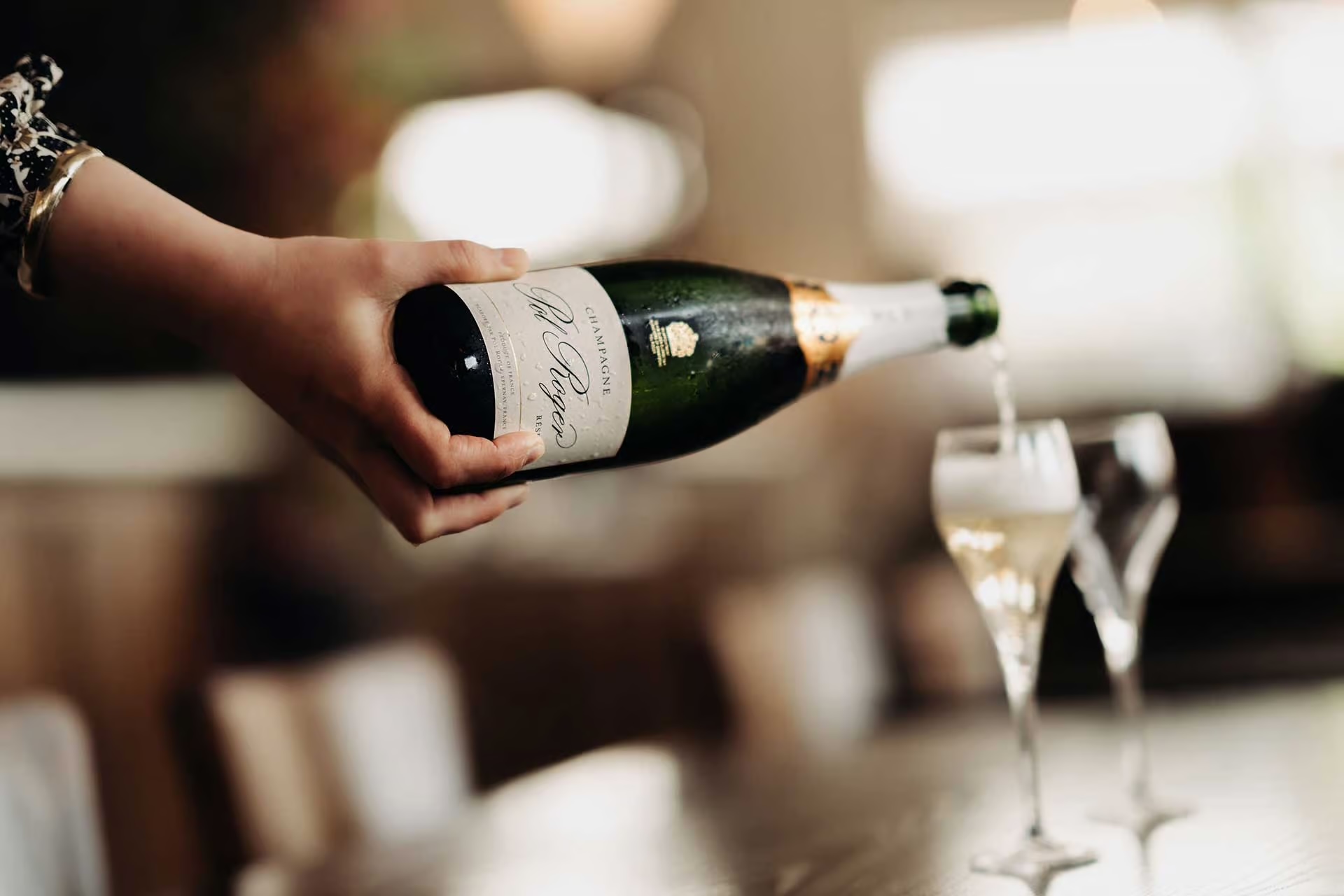 Hand pouring champagne from a bottle into a flute glass on a table.