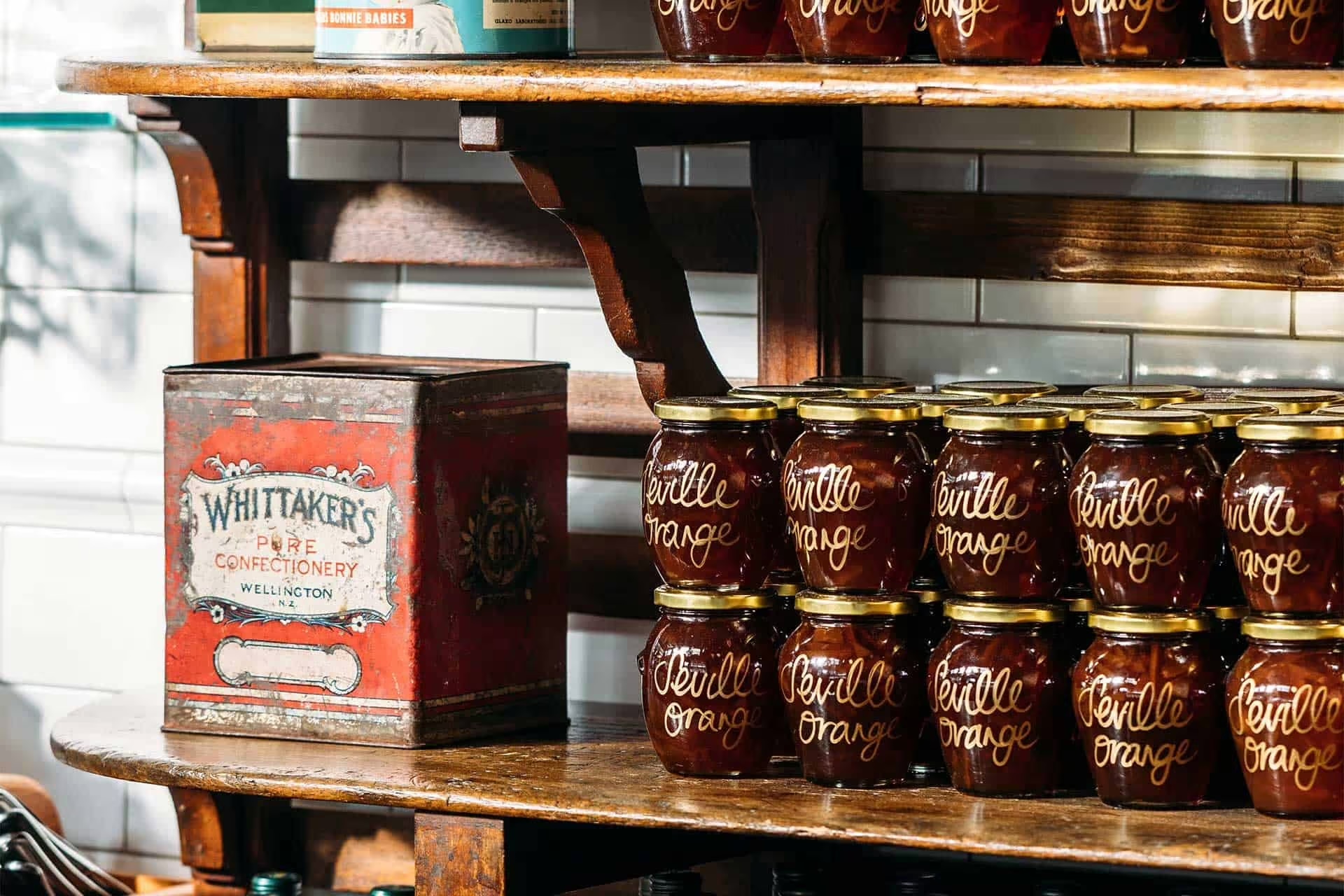 Wooden shelf displaying multiple jars of Seville orange marmalade next to a vintage red Whittaker's Pure Confectionery tin.