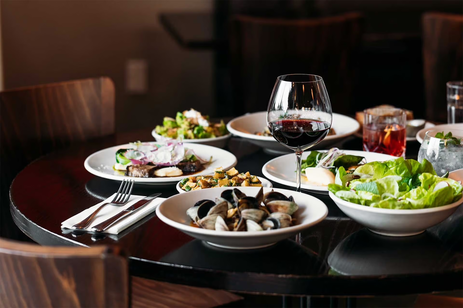 Dining table with plates of clams, salad, cooked vegetables, and sliced meat, accompanied by a glass of red wine and other drinks.