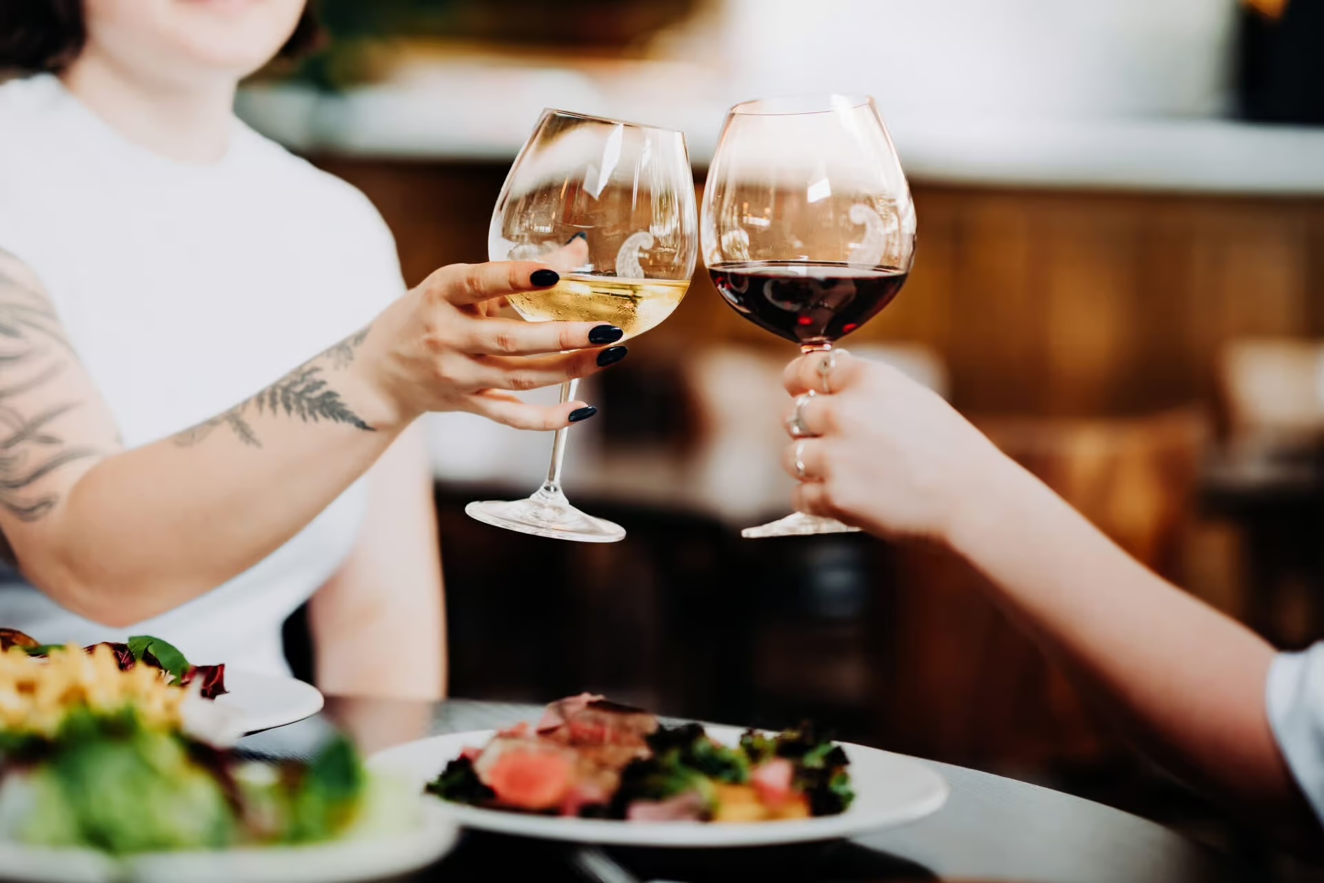 Two people clinking glasses of red and white wine over a table with plates of food.