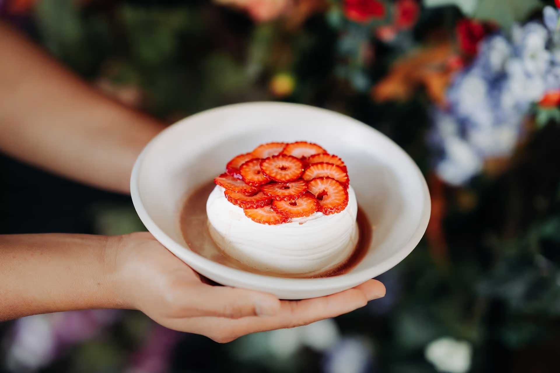 Hands holding a white dessert topped with sliced strawberries on a white plate.