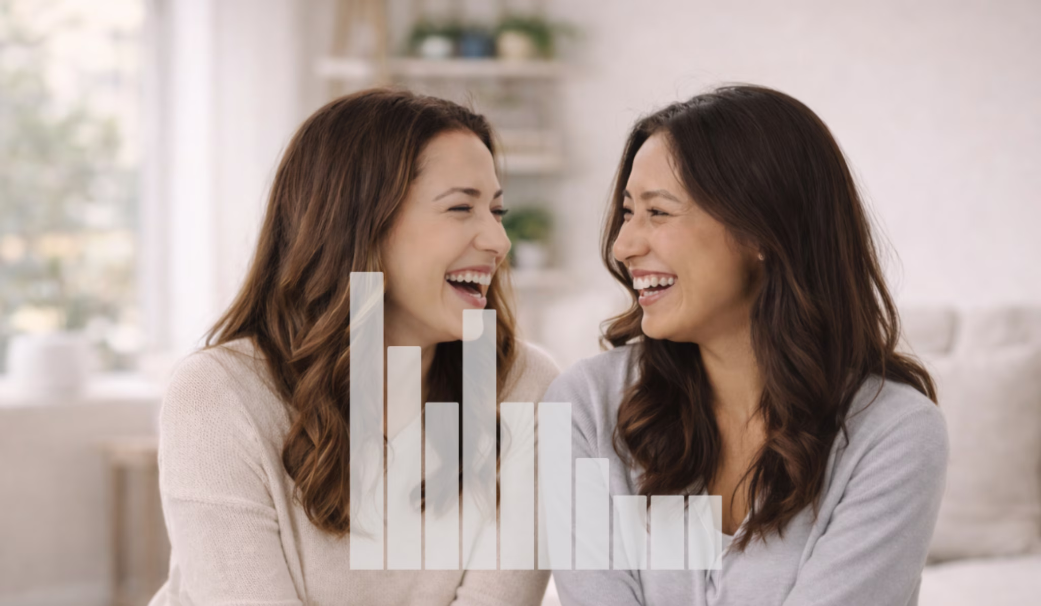 Two women with brown hair laughing joyfully while looking at each other indoors.