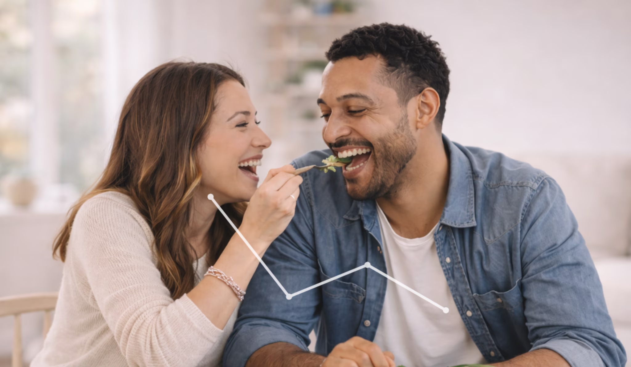 Happy woman feeding salad to smiling man in a bright kitchen.
