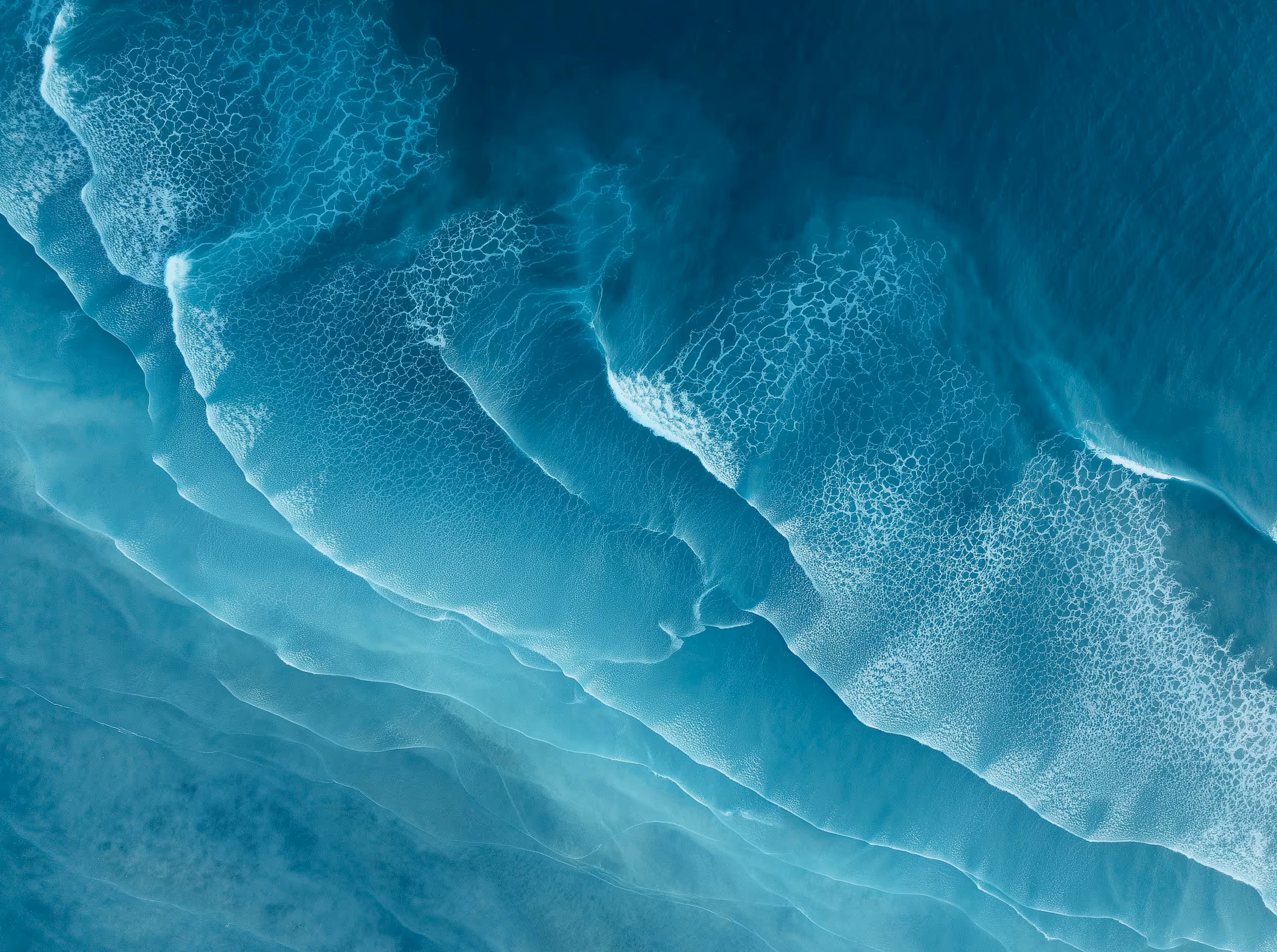 Aerial view of turquoise ocean waves gently rolling and foaming onto a sandy shore.