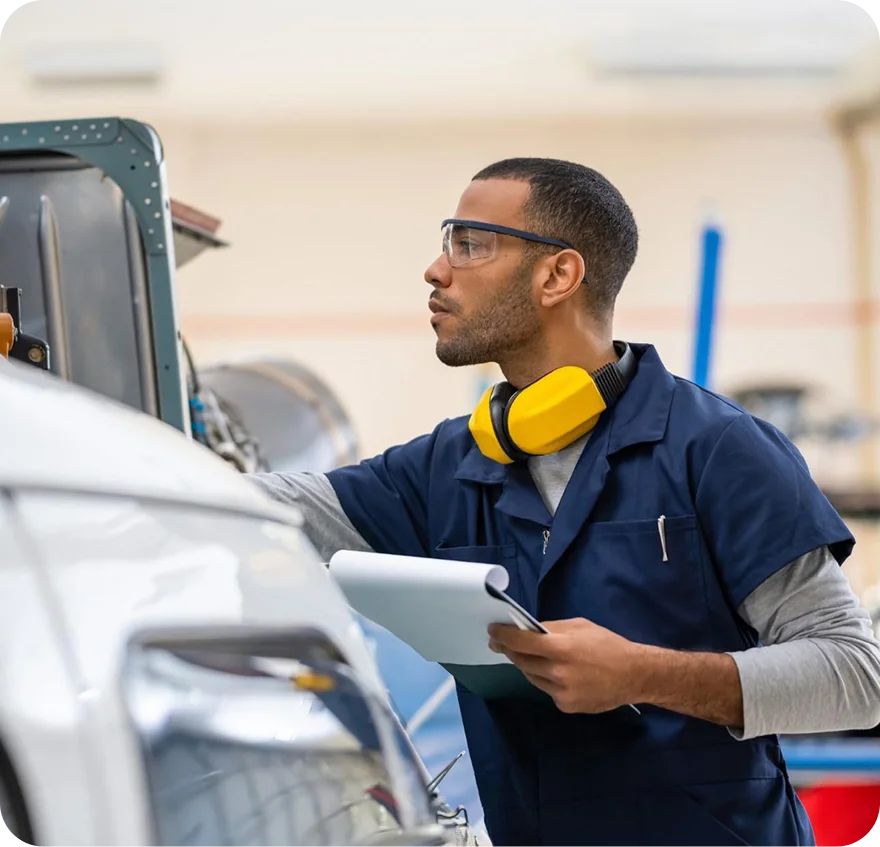 Mechanic wearing safety glasses and yellow ear protectors around neck inspecting a vehicle with clipboard in hand.