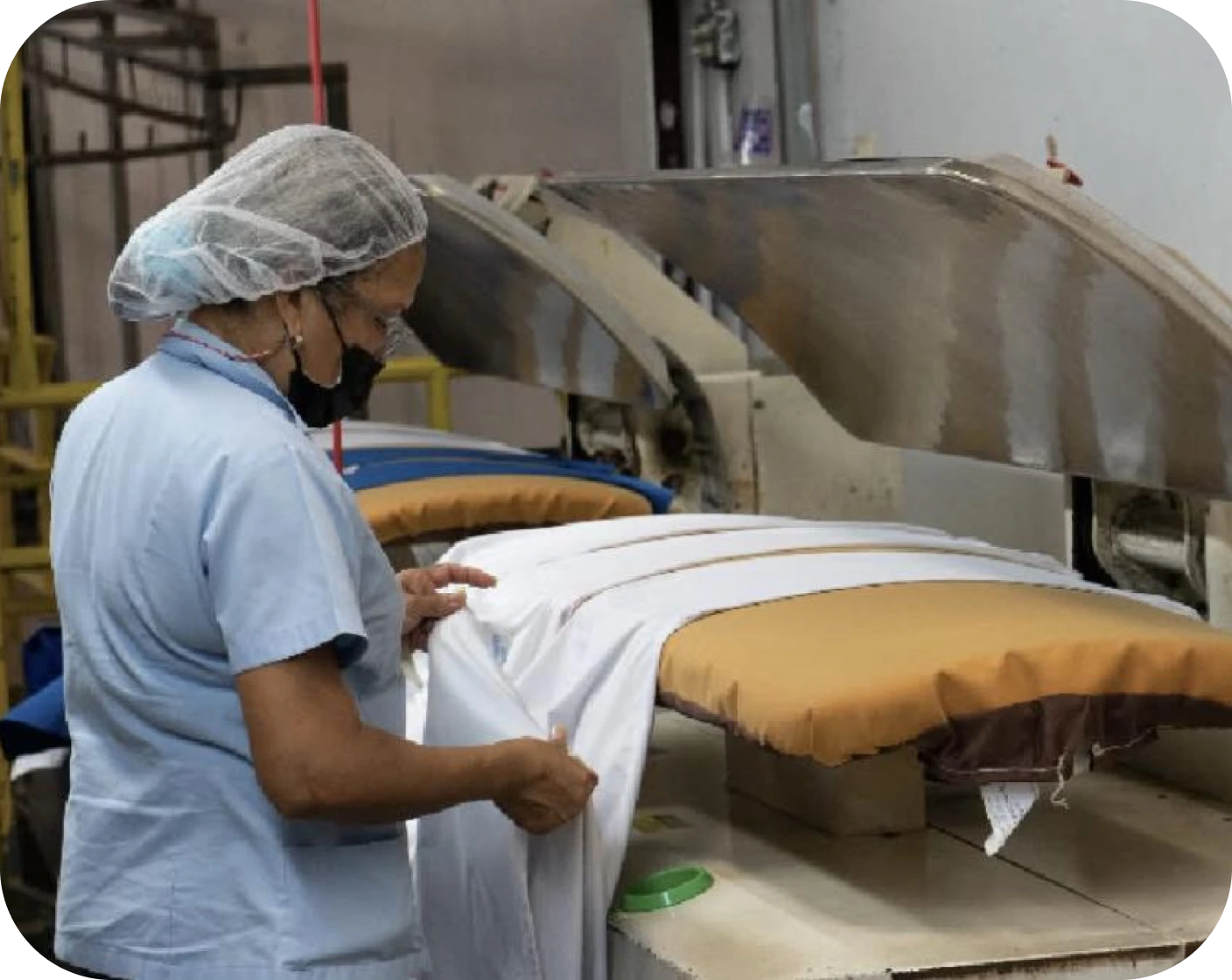 Worker wearing a hairnet and mask handling white fabric on an ironing press machine in a laundry or manufacturing setting.