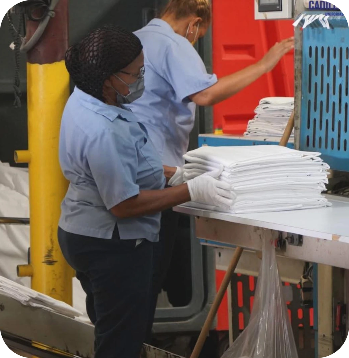 Two workers in blue uniforms folding and stacking white linens on a table in a laundry facility.