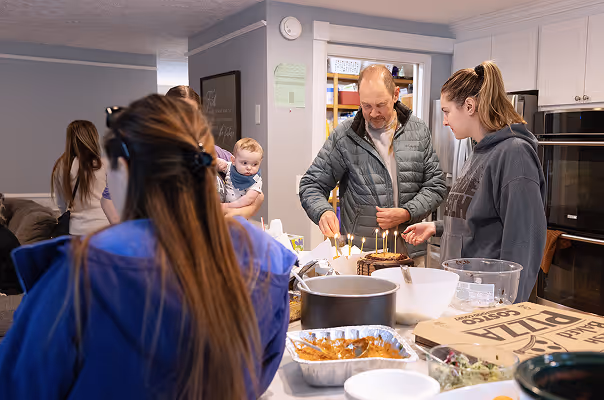 A small group gathered in a kitchen where a man and woman are lighting candles on a cake, with food and pizza boxes on the counter.