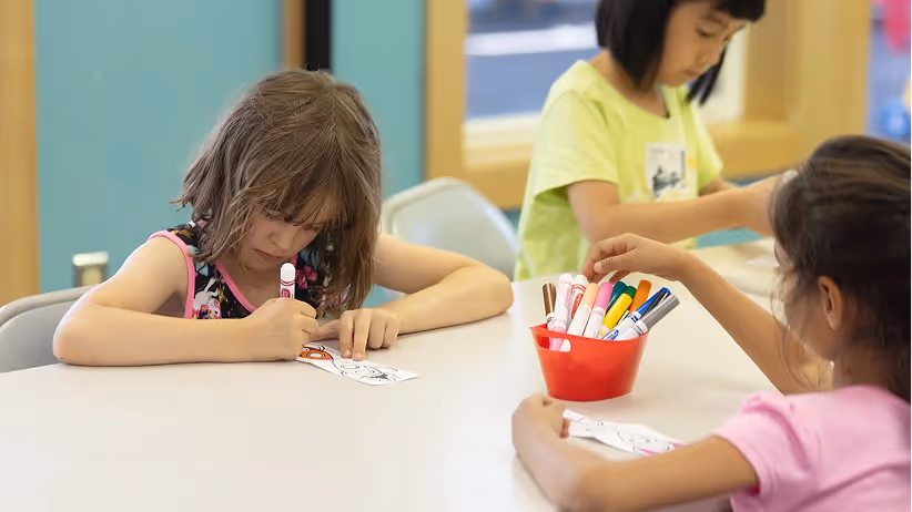 Three children sitting at a table, coloring on paper with markers, focused on their artwork.