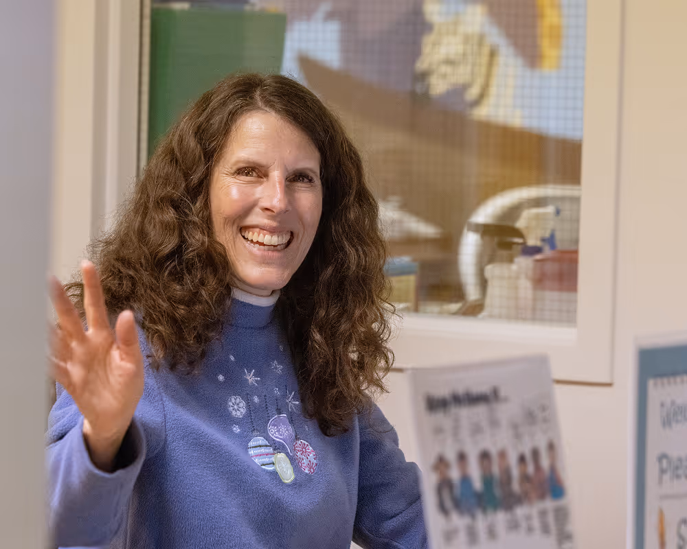 Smiling woman with curly brown hair wearing a blue sweater with holiday ornament designs, waving her hand indoors.