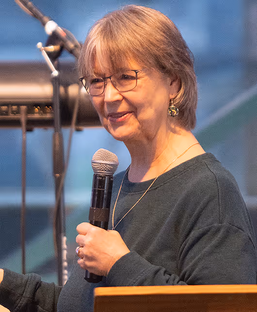 Smiling older woman with short hair and glasses speaking into a microphone on stage during a service.