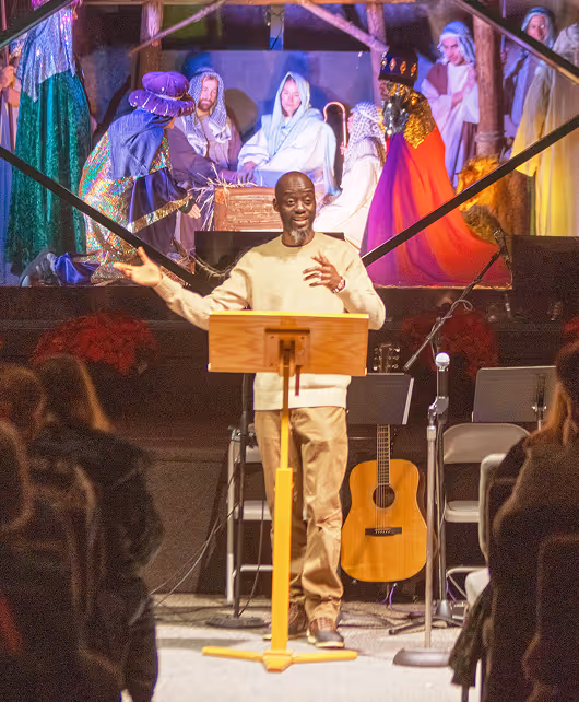 Pastor Mark Nsimbi speaking at a wooden podium with a nativity scene backdrop and seated audience at Red-Wood Falls Church.