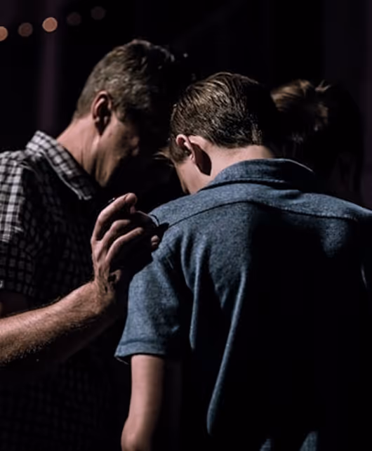 Two people holding hands in a dimly lit setting, in a moment of prayer and reflection.