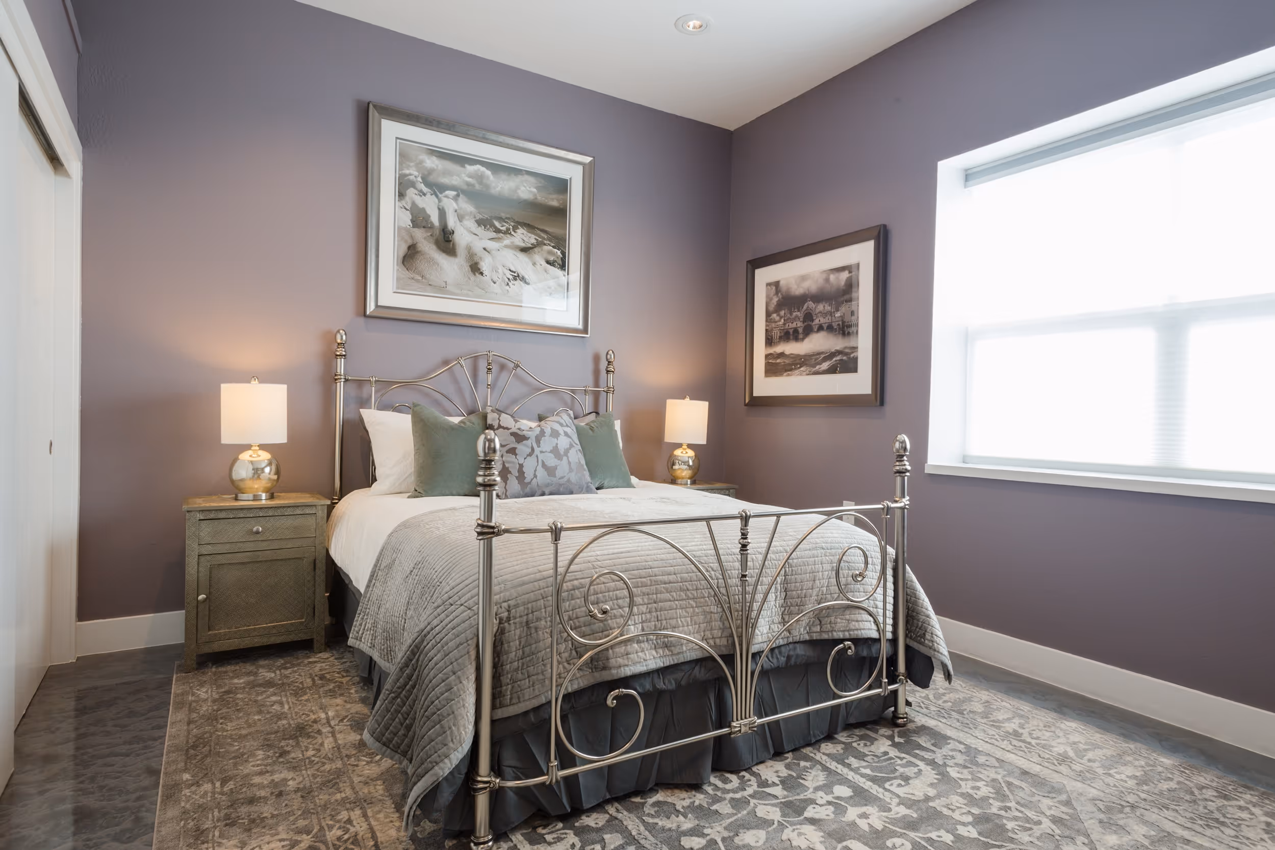 Bedroom with a metal frame bed, nightstands with lamps, and framed black-and-white photos on lavender walls.