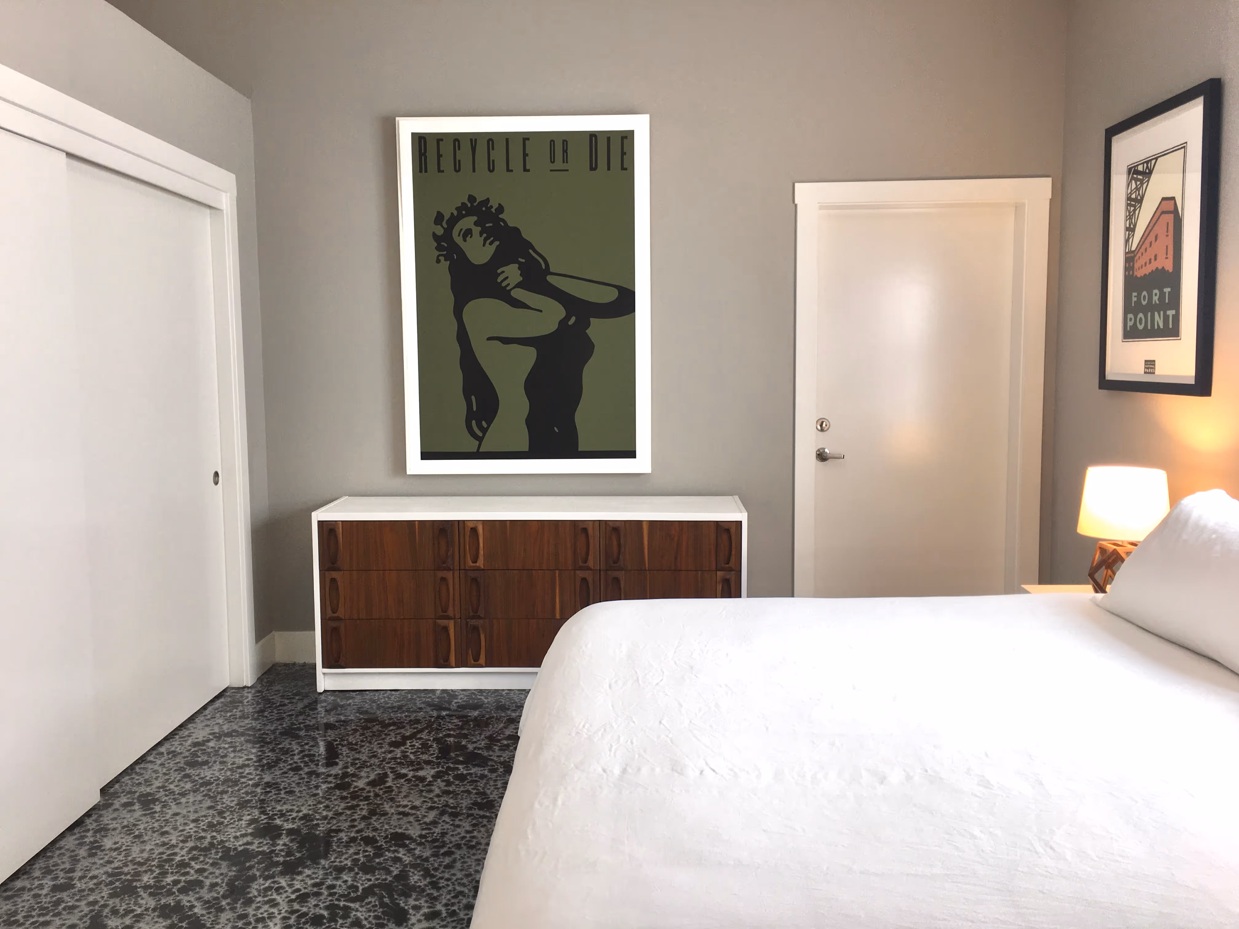 Minimalist bedroom with white bed, wooden dresser, and two framed posters on gray walls.