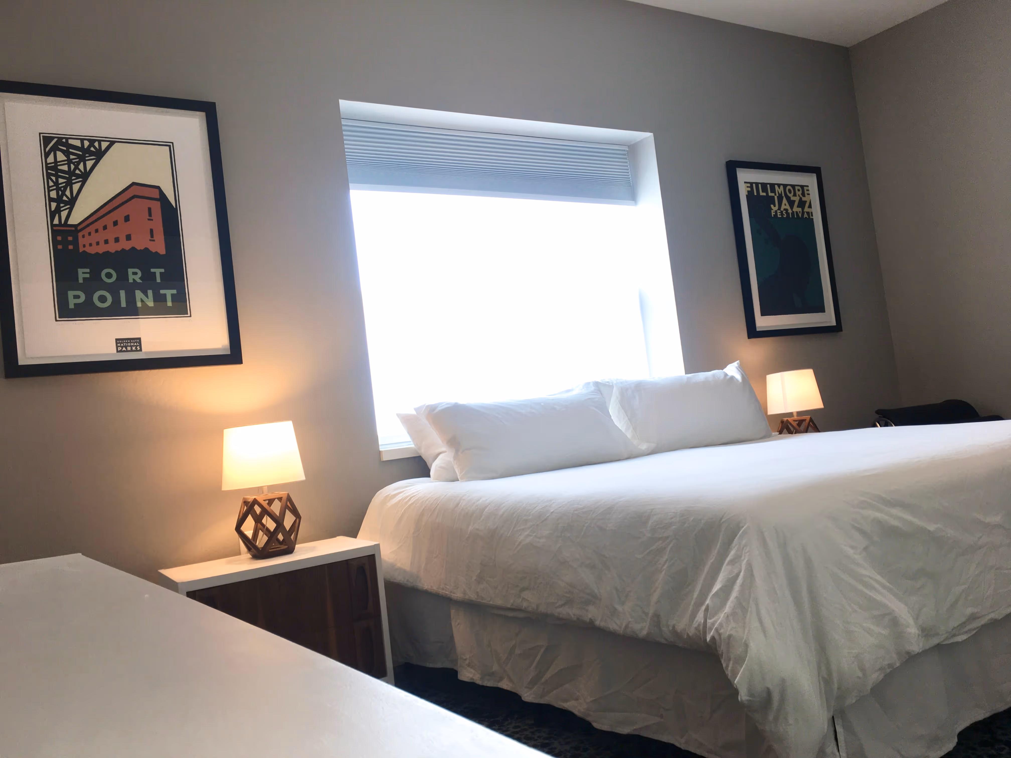 Minimalist bedroom with a large king-sized bed, white bedding, two bedside lamps, and framed posters on gray walls.