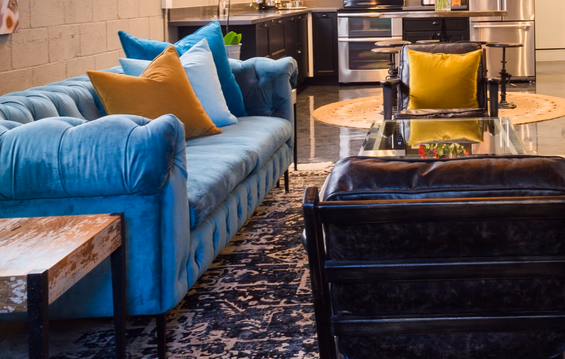 Blue velvet sofa with multiple throw pillows next to black leather chairs around a glass coffee table in a living room with a kitchen in the background.