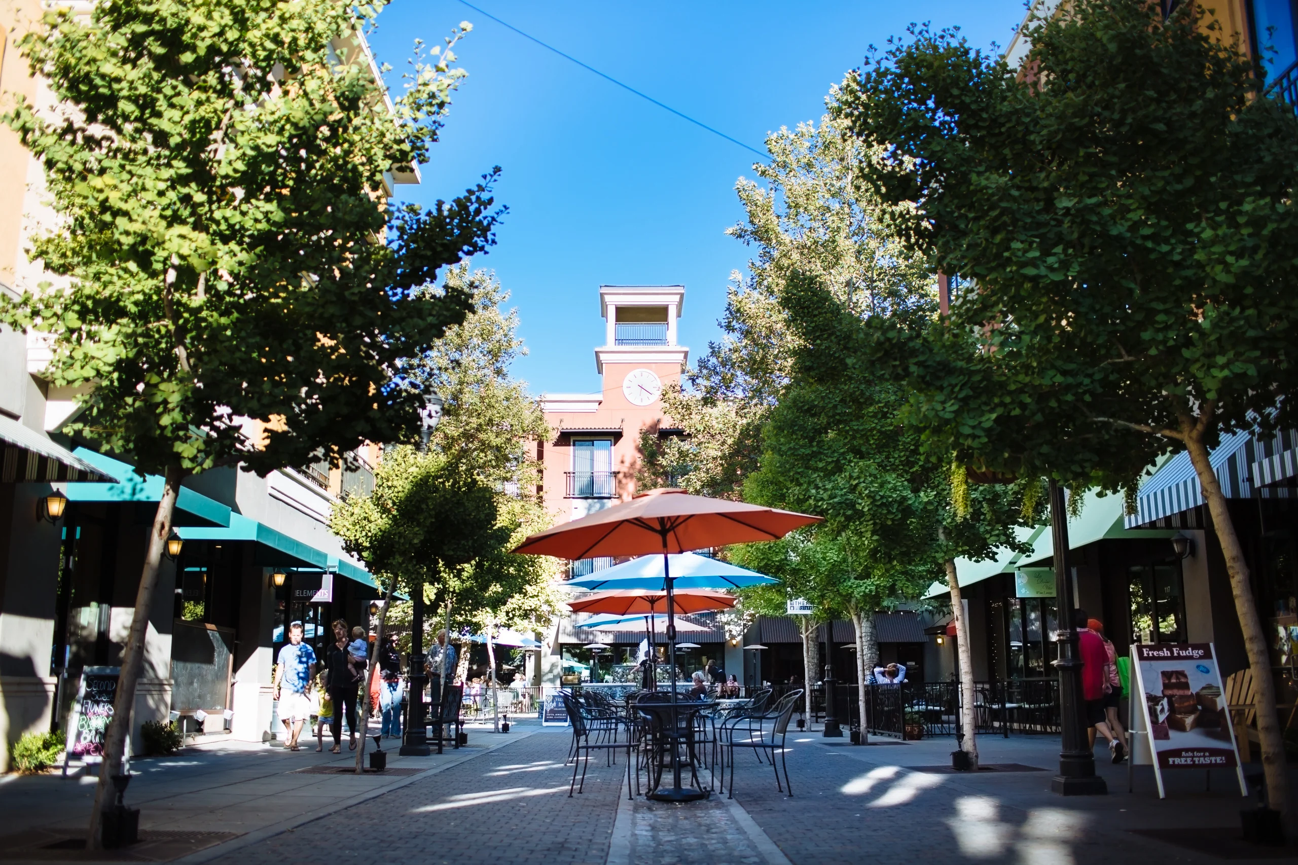 Outdoor urban plaza with tables, umbrellas, trees, and people walking near shops under clear blue sky.