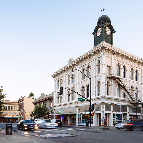 Historic white three-story building with a clock tower at a busy street corner with cars and pedestrians.