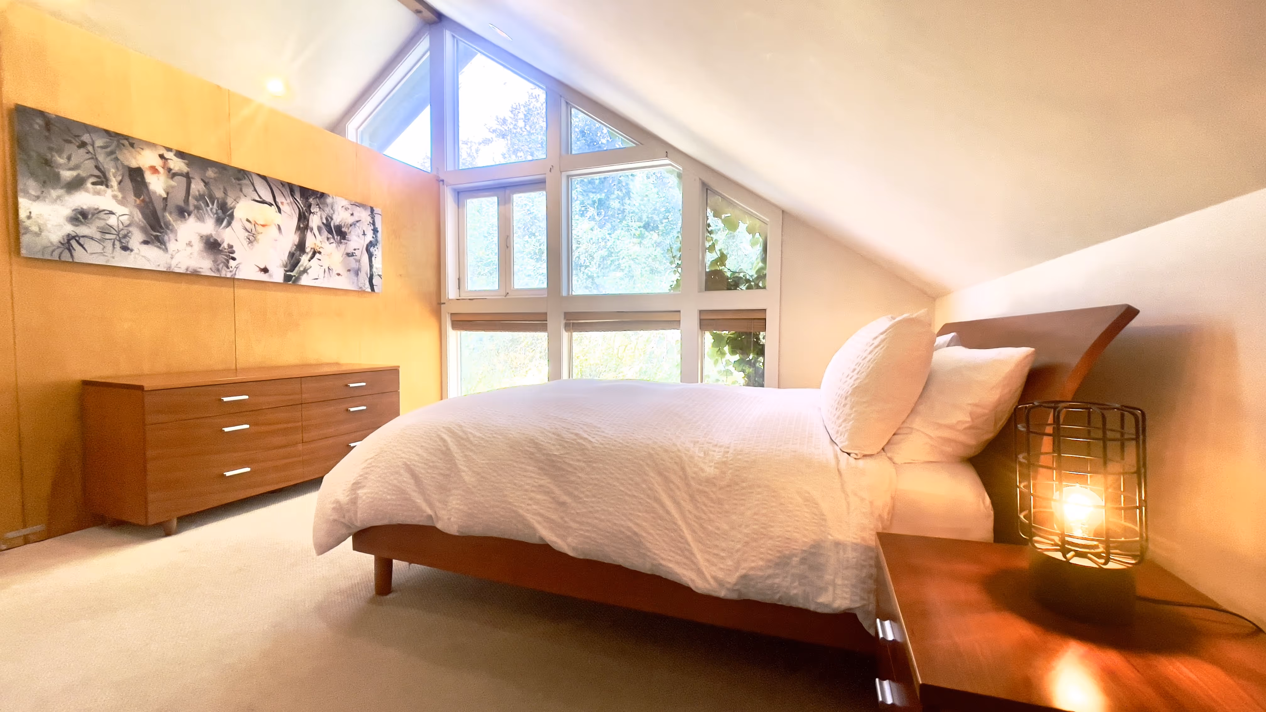 Bright bedroom with a wooden bed, white bedding, large triangular window, wooden dresser, and a lit lamp on the nightstand.