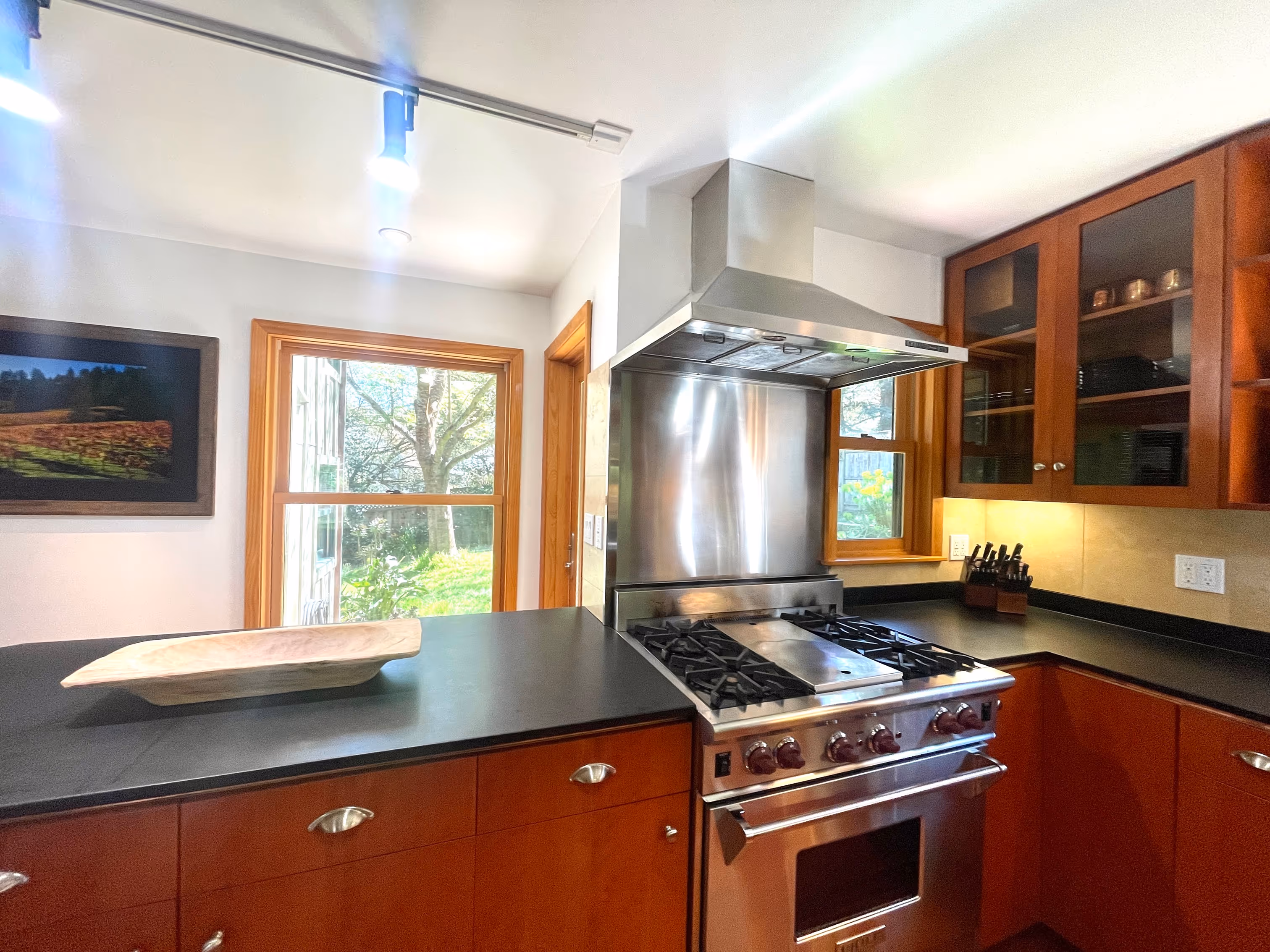 Modern kitchen with stainless steel gas stove and hood, dark countertops, wooden cabinets, and a window showing trees outside.