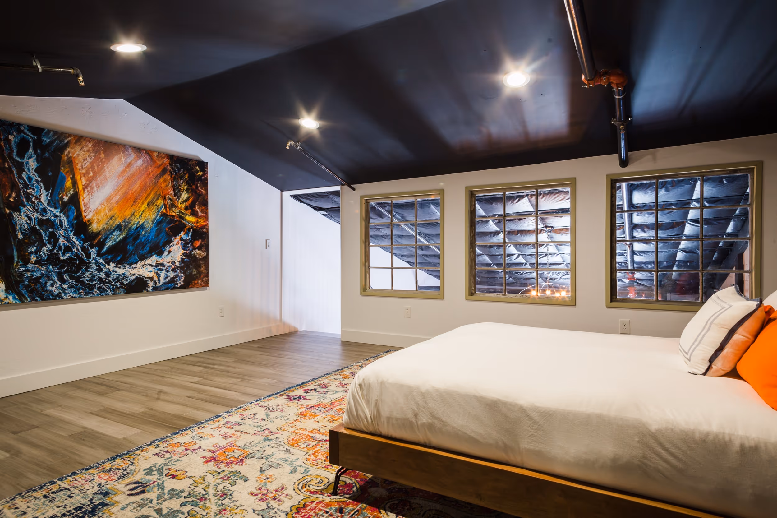 Bedroom with a white bed, colorful rug, three large windows showing an insulated attic ceiling, black ceiling, and abstract painting on wall.