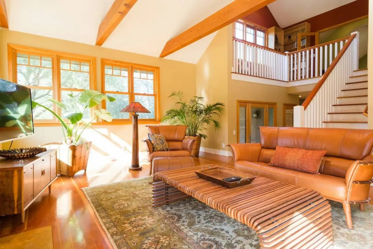 Bright living room with tan leather sofa and armchair, wooden coffee table, large green plants, and tall windows.