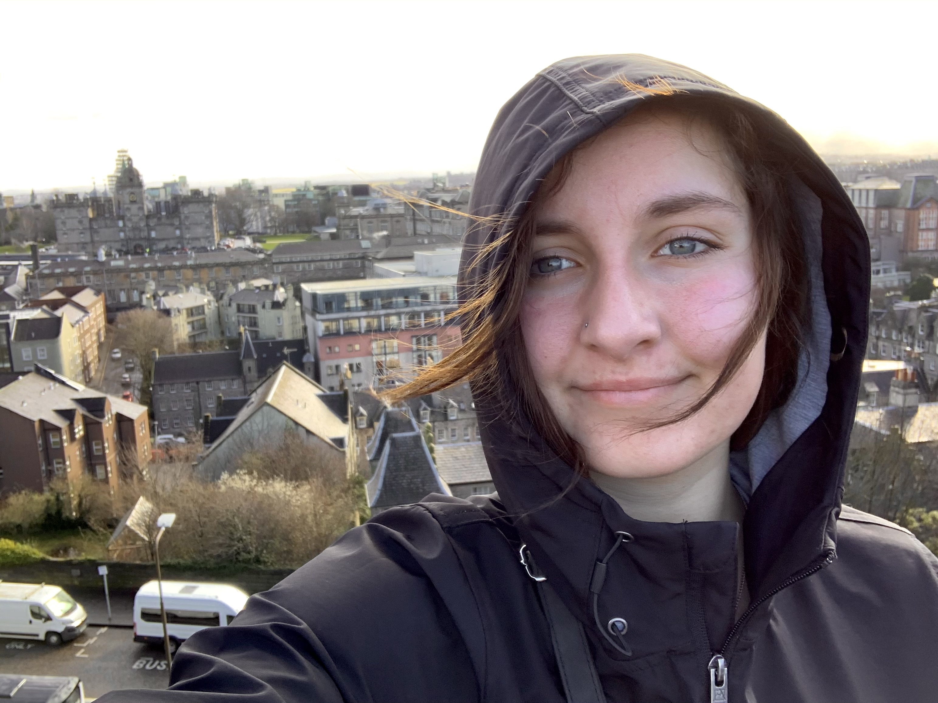 Young woman in a black hooded jacket smiling with city buildings and clear sky in the background.