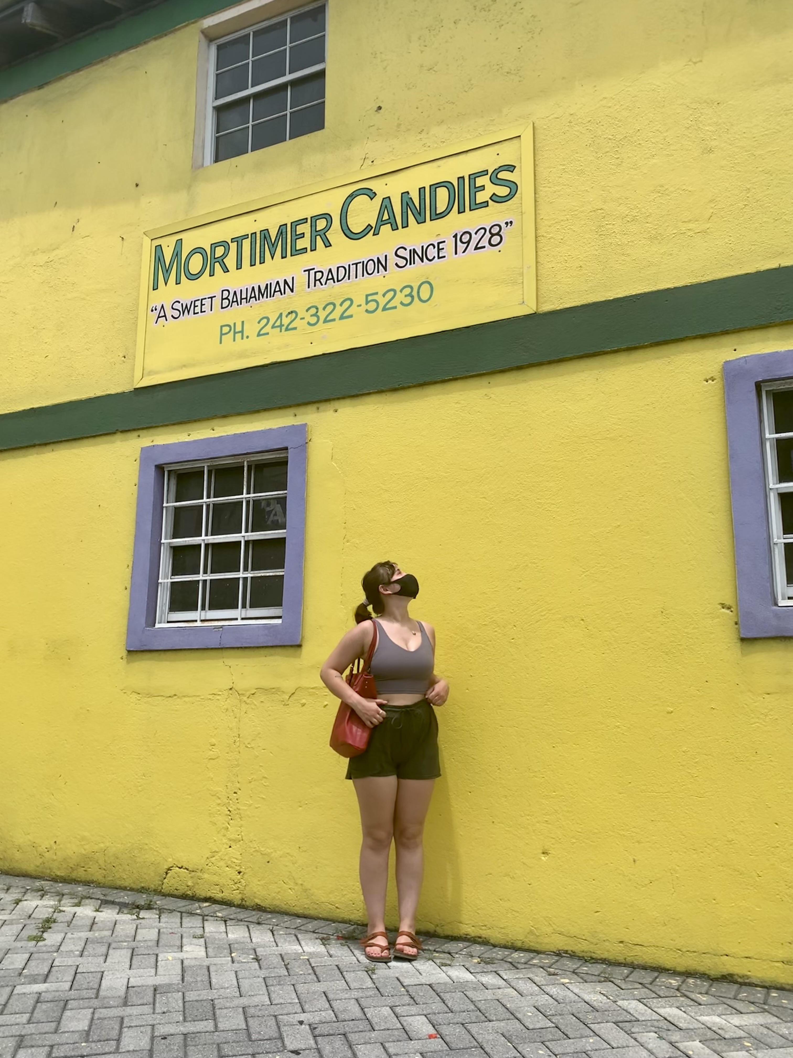 Woman wearing mask stands against bright yellow wall beneath Mortimer Candies sign with phone number.