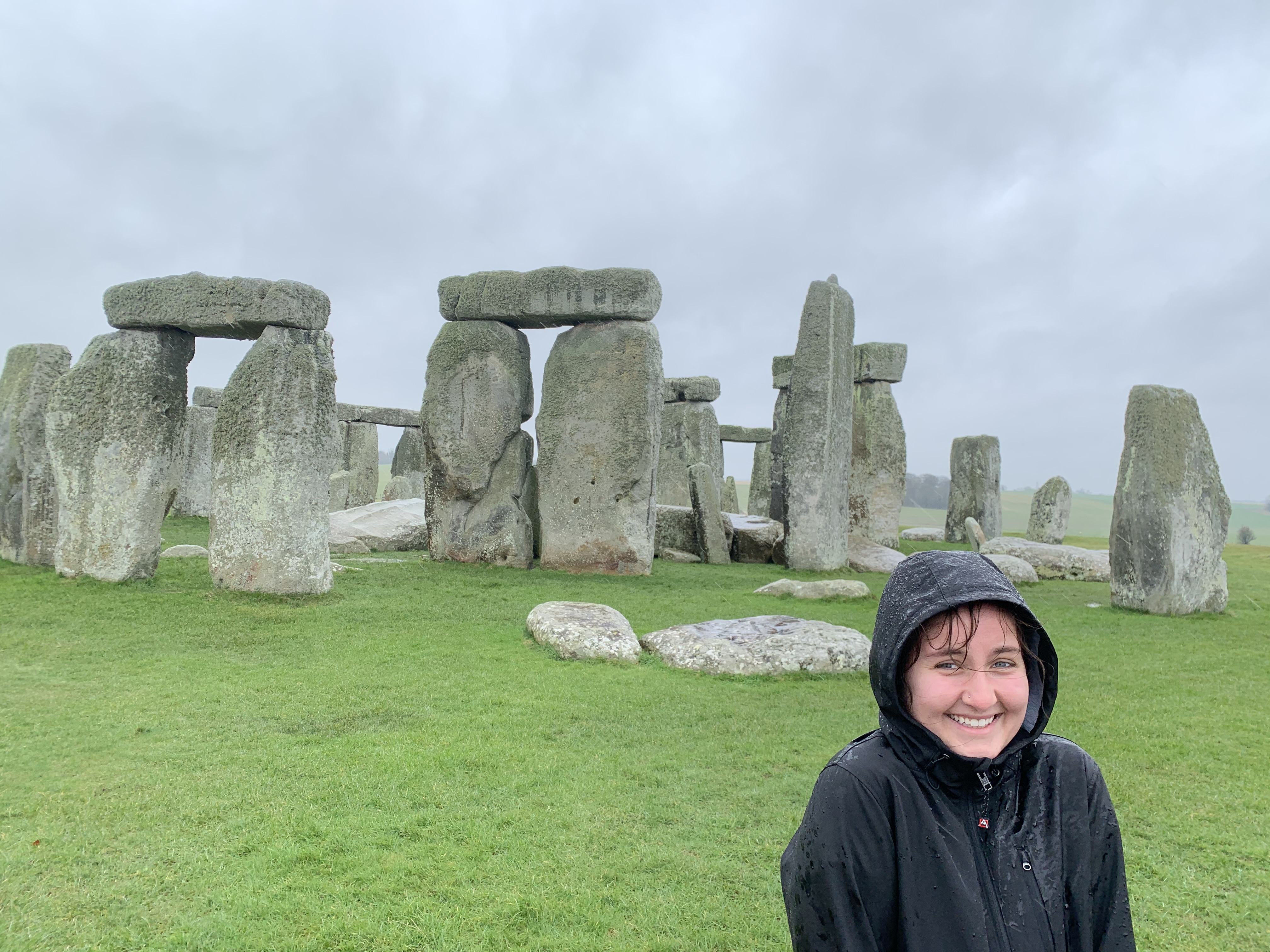 Smiling person in a black raincoat stands on green grass in front of Stonehenge under a cloudy sky.