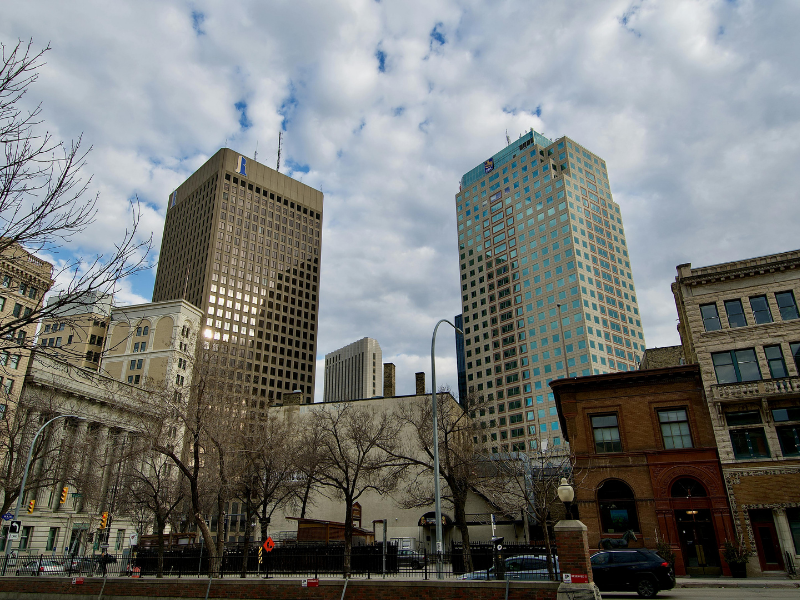 Urban cityscape with leafless trees, historic buildings, and two tall modern office towers under a cloudy sky.