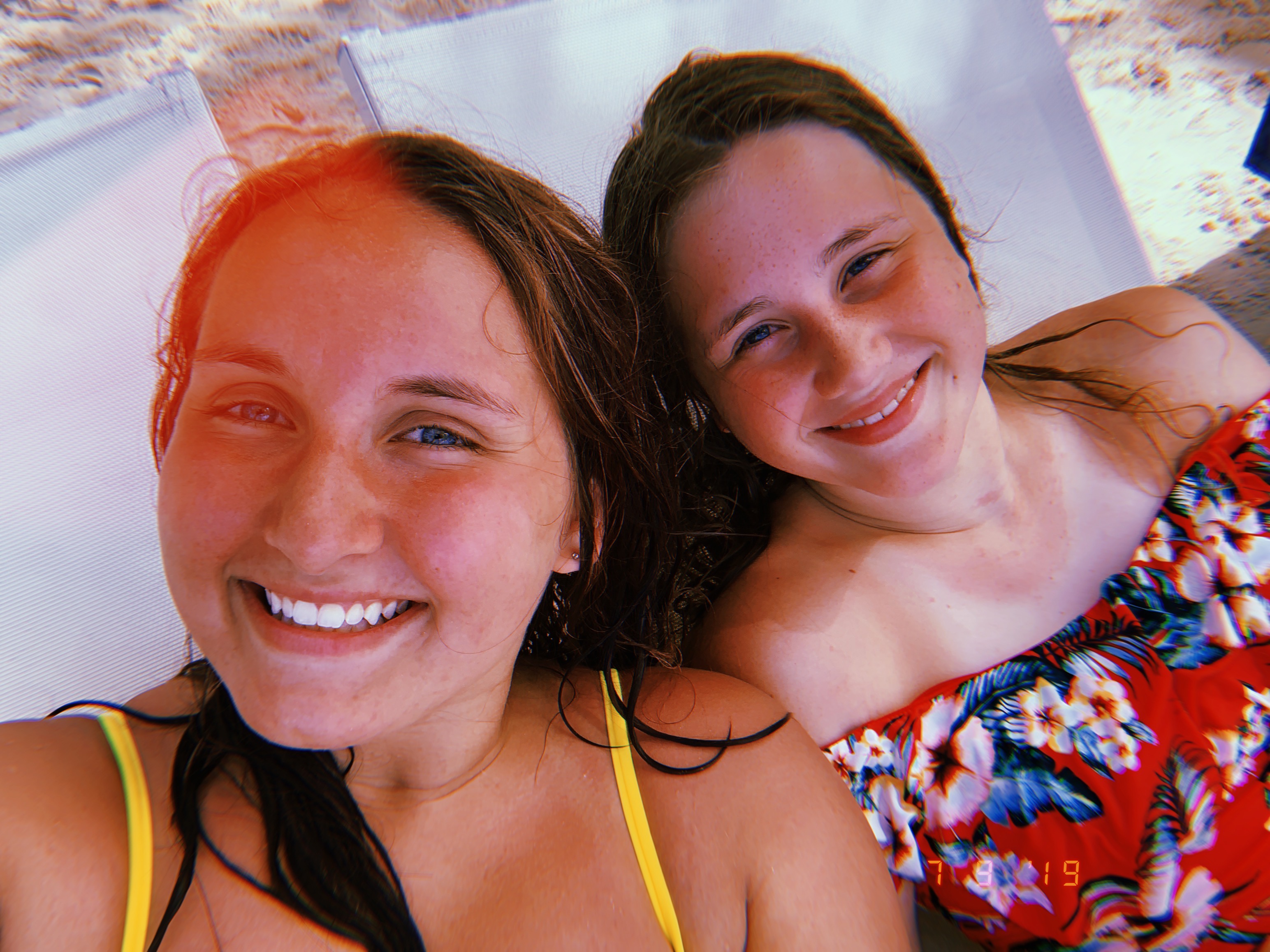 Two smiling women lying on beach loungers, one in a yellow swimsuit and the other in a floral top.