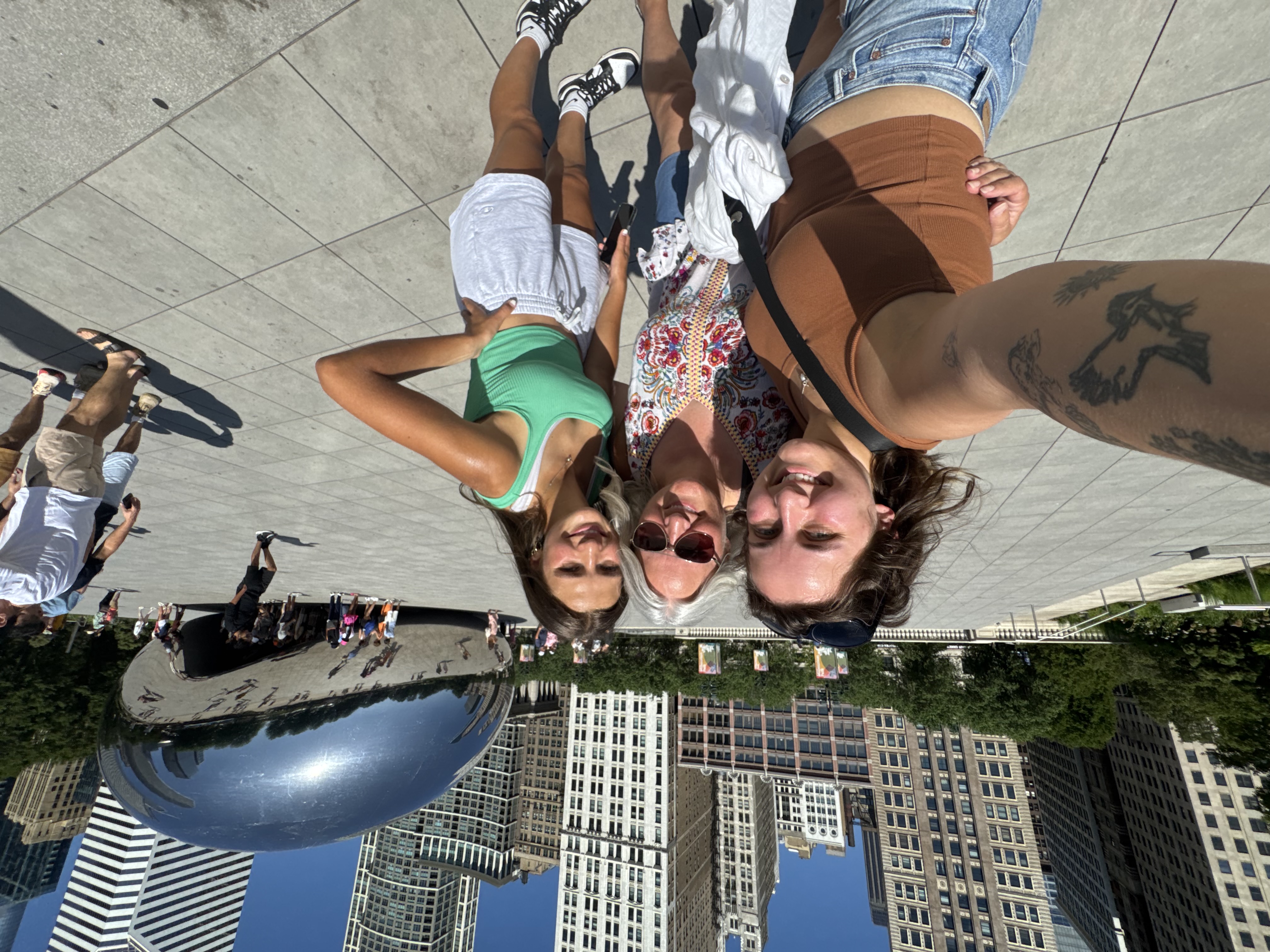 Three women smiling and taking a selfie in front of Chicago's Cloud Gate sculpture on a sunny day.