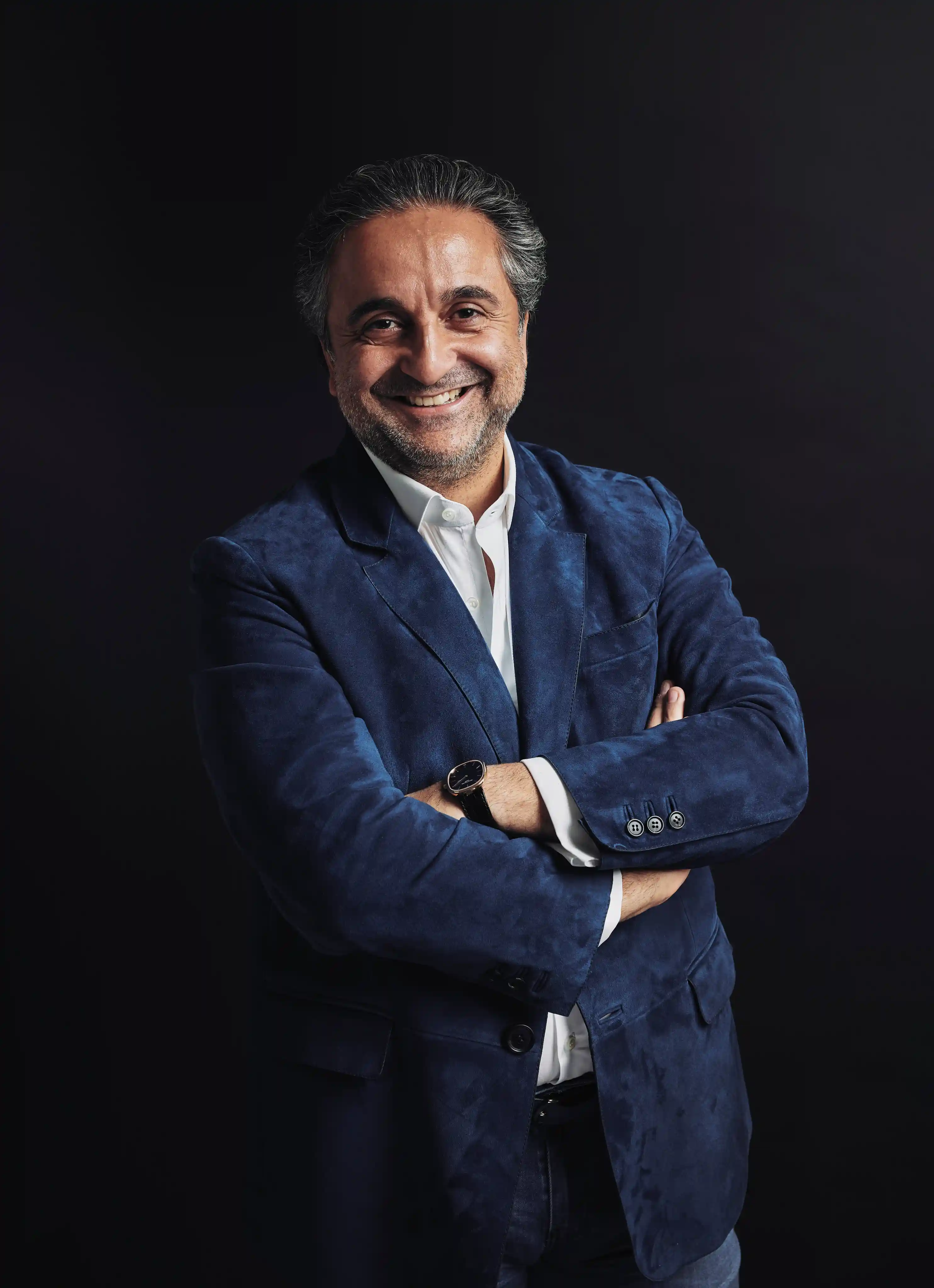 Homme souriant bras croisés portant une veste bleu marine et une chemise blanche sur fond noir.