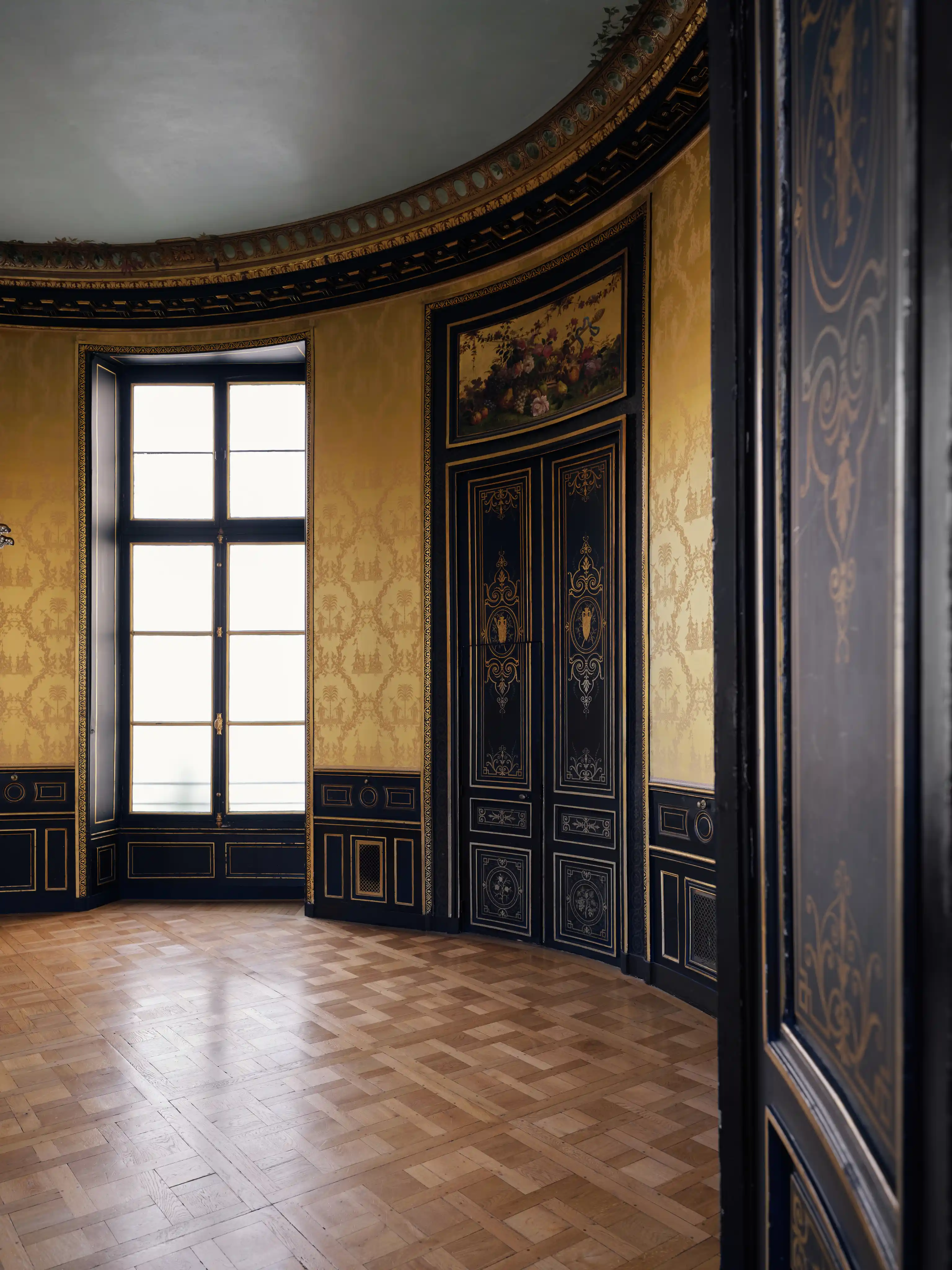 Salle vide avec parquet en bois et murs décorés de panneaux noirs avec dorures et une fenêtre haute laissant entrer la lumière naturelle.