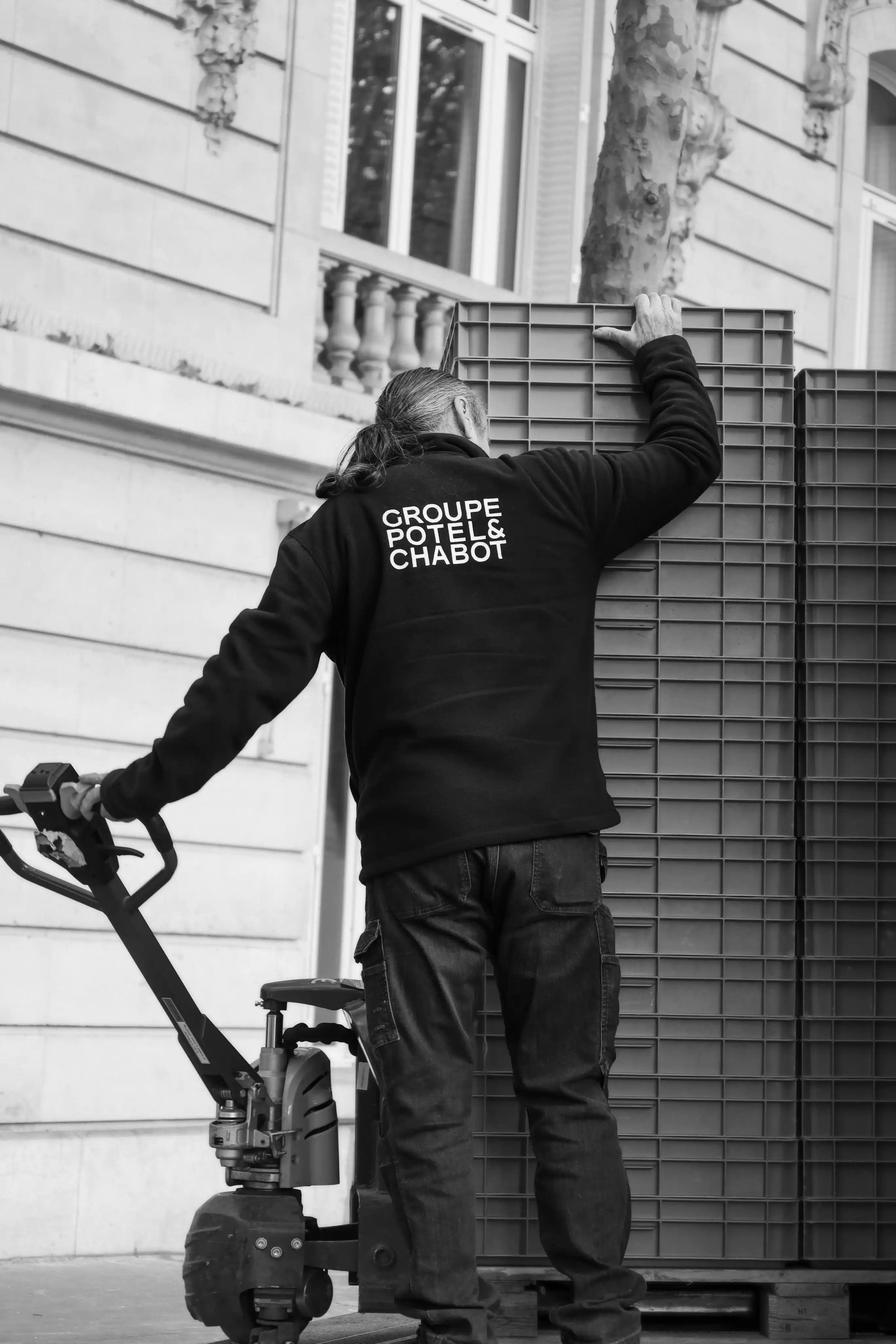 Un homme en veste noire avec le logo Groupe Potel & Chabot déplace des caisses empilées à l'aide d'un transpalette devant un bâtiment en pierre.
