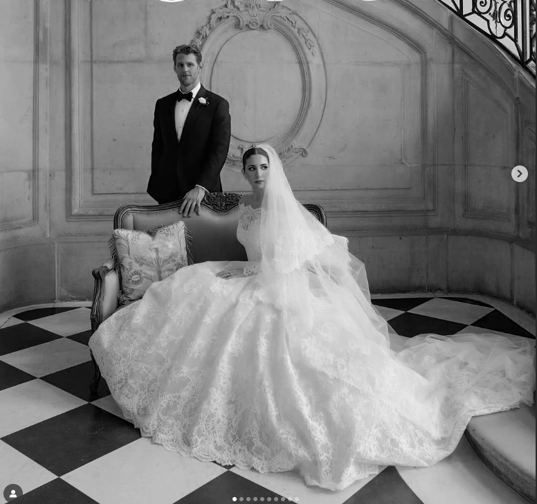 Mariée en robe longue en dentelle assise sur un canapé devant un mur orné, avec le marié en costume noir debout derrière elle sur un sol à damier.