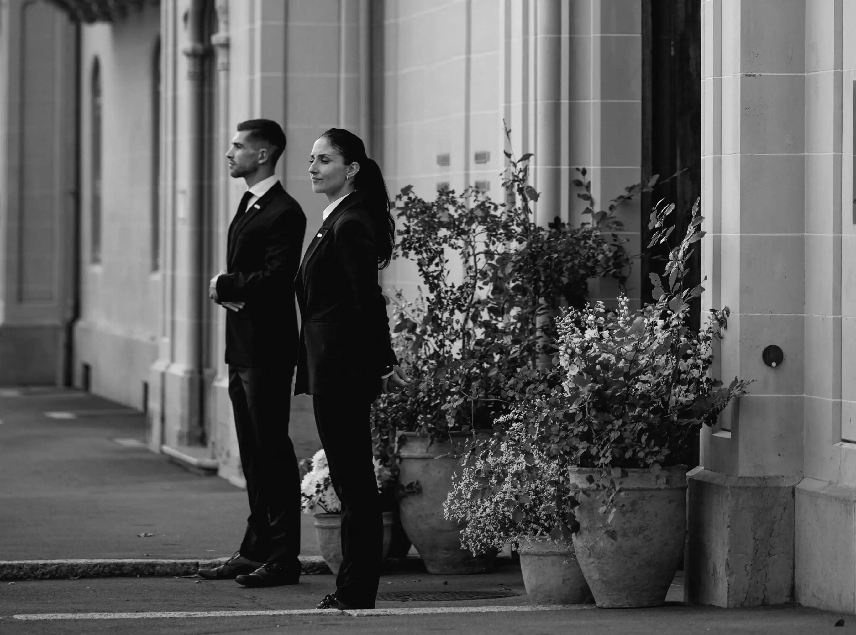 Deux agents de sécurité en costume sombre debout devant un bâtiment aux murs en pierre, à côté de grands pots de fleurs.