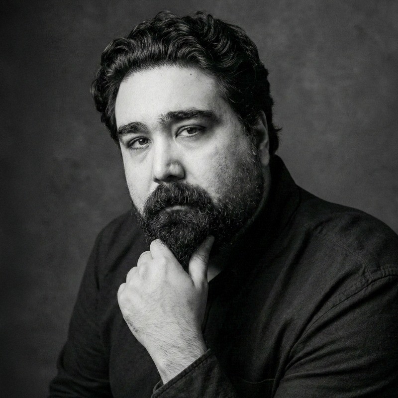Black and white portrait of Storm Brewer, a man with curly hair and a beard resting his chin on his hand and looking thoughtfully at the camera.