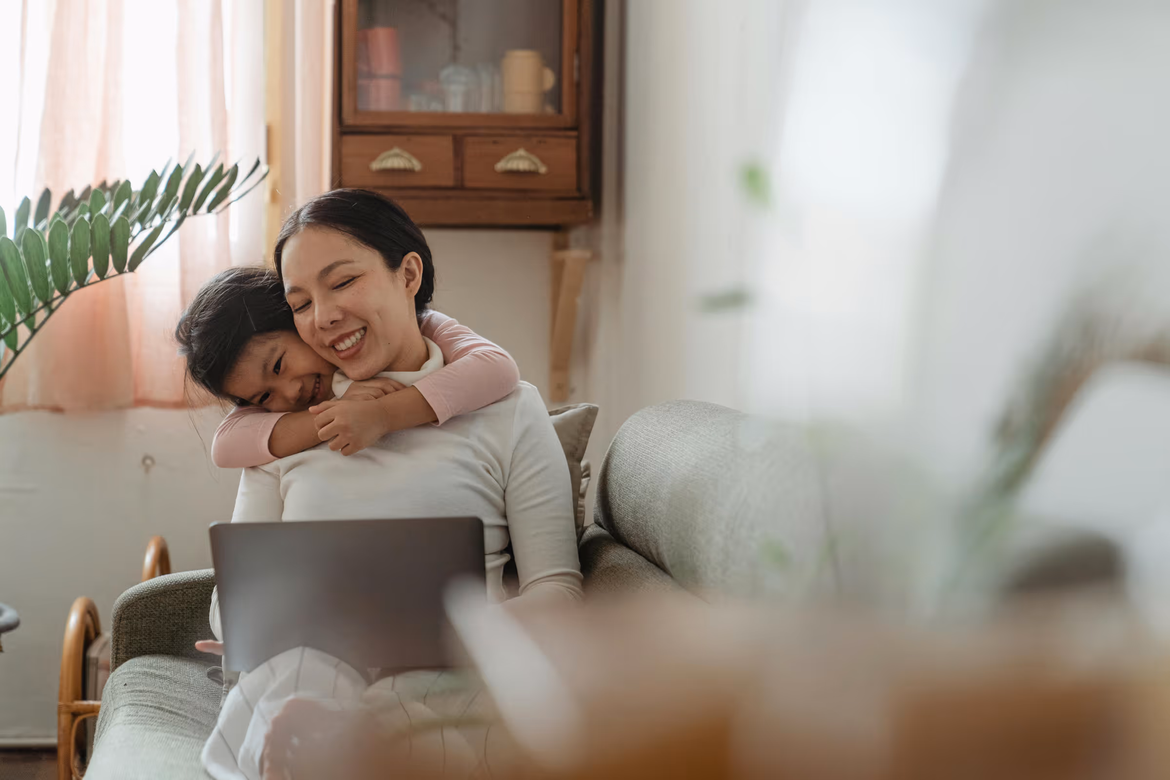 Smiling woman sitting on a couch with a laptop while a child hugs her from behind.