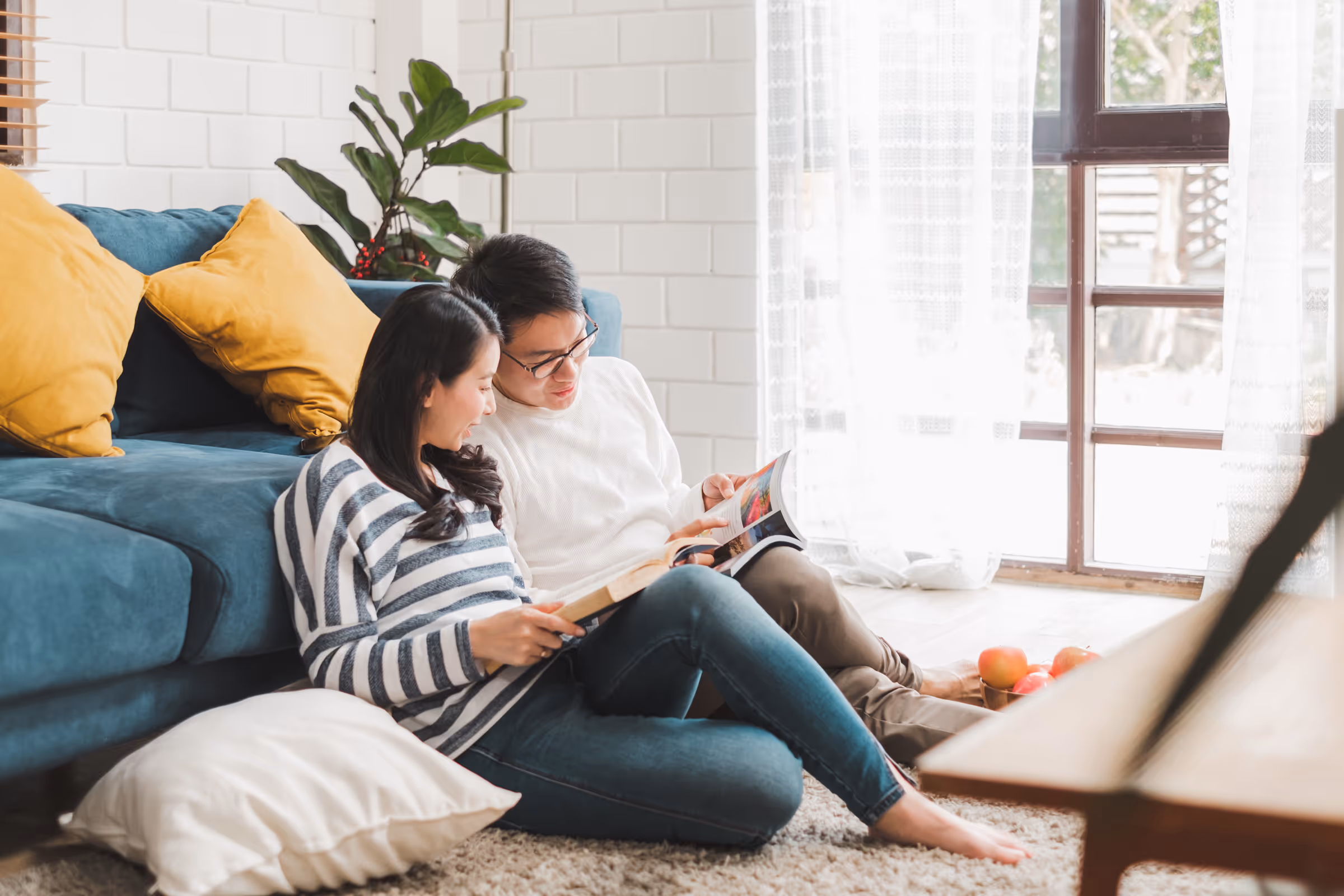Couple sitting on carpeted floor leaning against blue couch with yellow pillows, reading books near large window with sheer curtains.